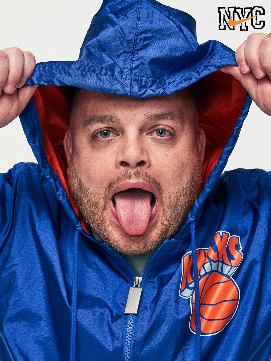 A man wearing a blue New York Knicks jacket and hood, holding the hood with both hands, sticking out his tongue, and looking directly at the camera.