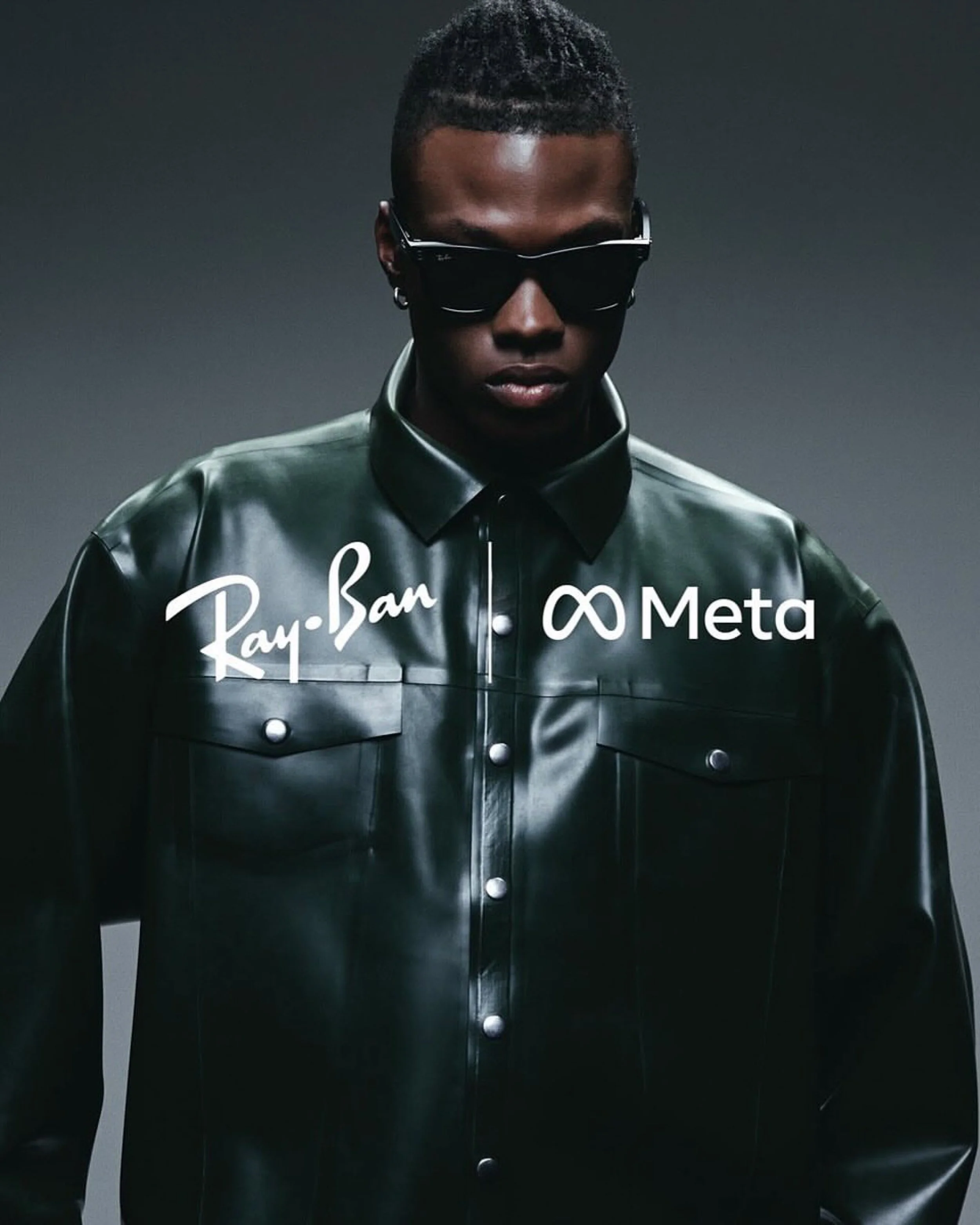 A person wearing black Ray-Ban sunglasses and a shiny black leather jacket with chrome buttons, standing against a dark background.