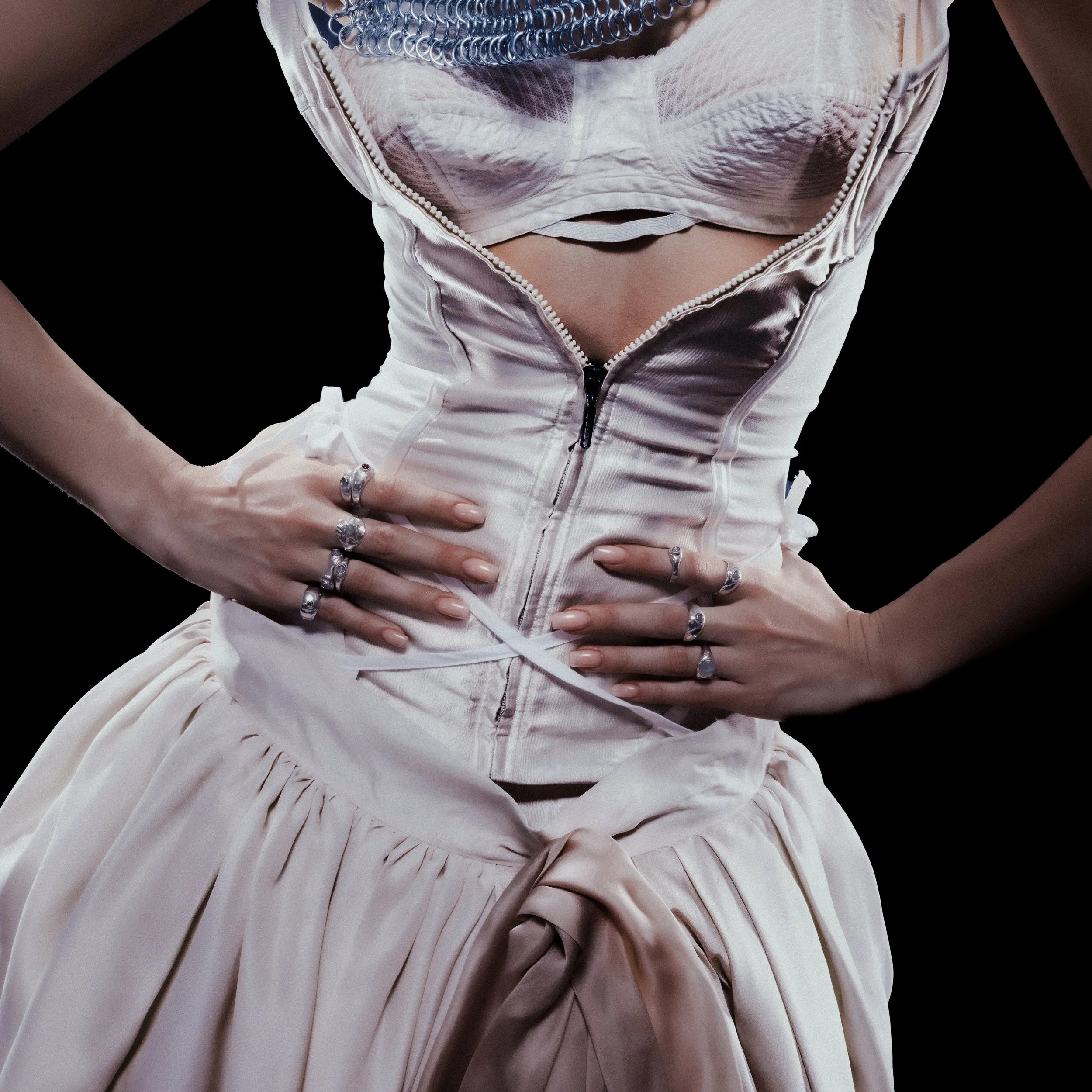 Close-up of a person's torso wearing a light pink or beige dress with a zipper, showing a layered undergarment or corset underneath. The person has multiple rings and bracelets on their hands, which are placed on their waist.