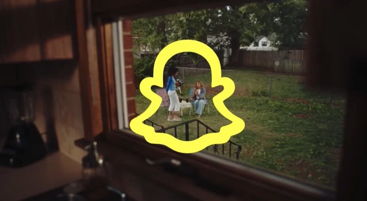 View through a kitchen window of two women in the backyard, one standing and the other sitting on a chair, with a Snapchat logo overlay.