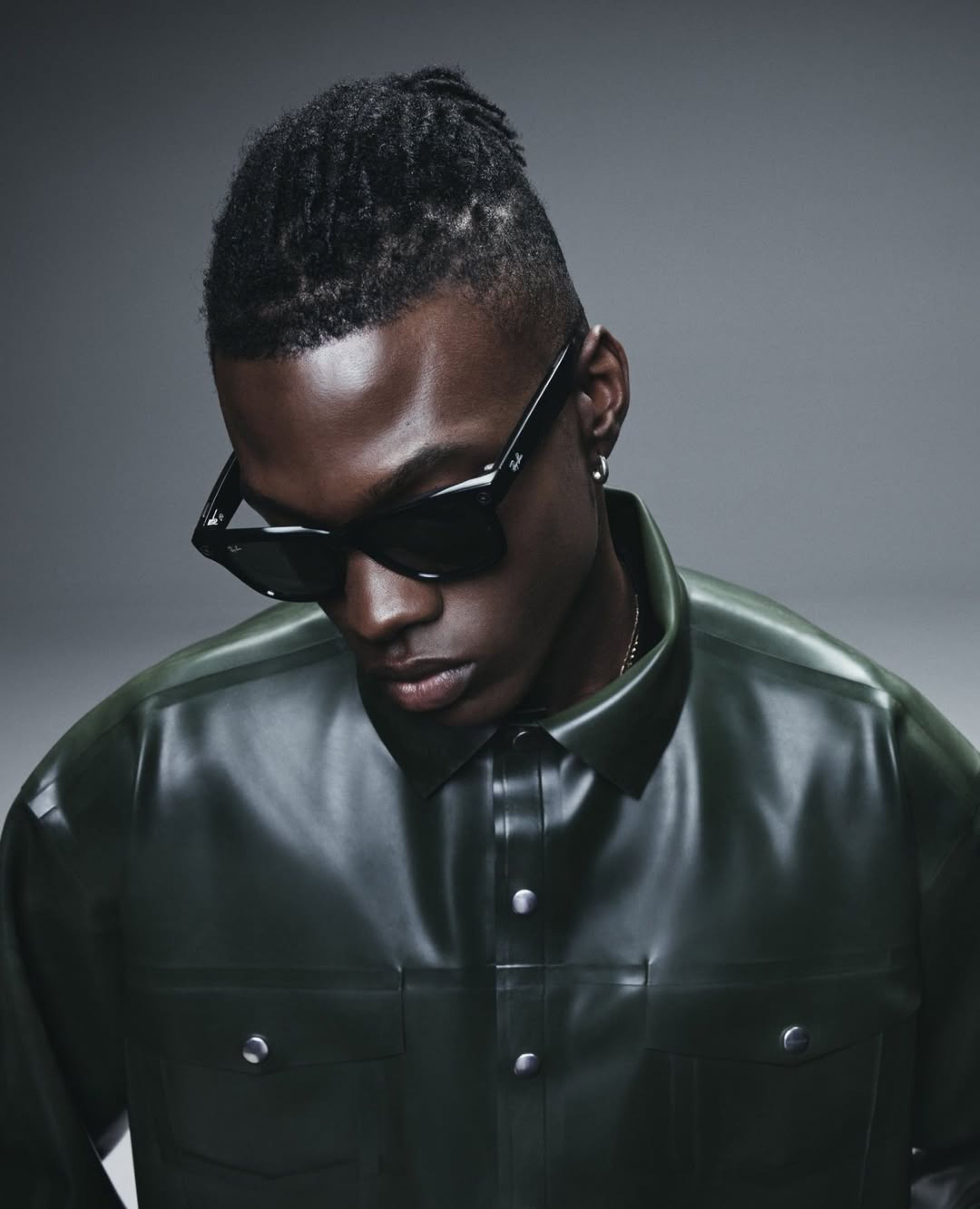 A person with short curly hair, wearing black sunglasses and a green leather jacket, appearing to be in a dark room.