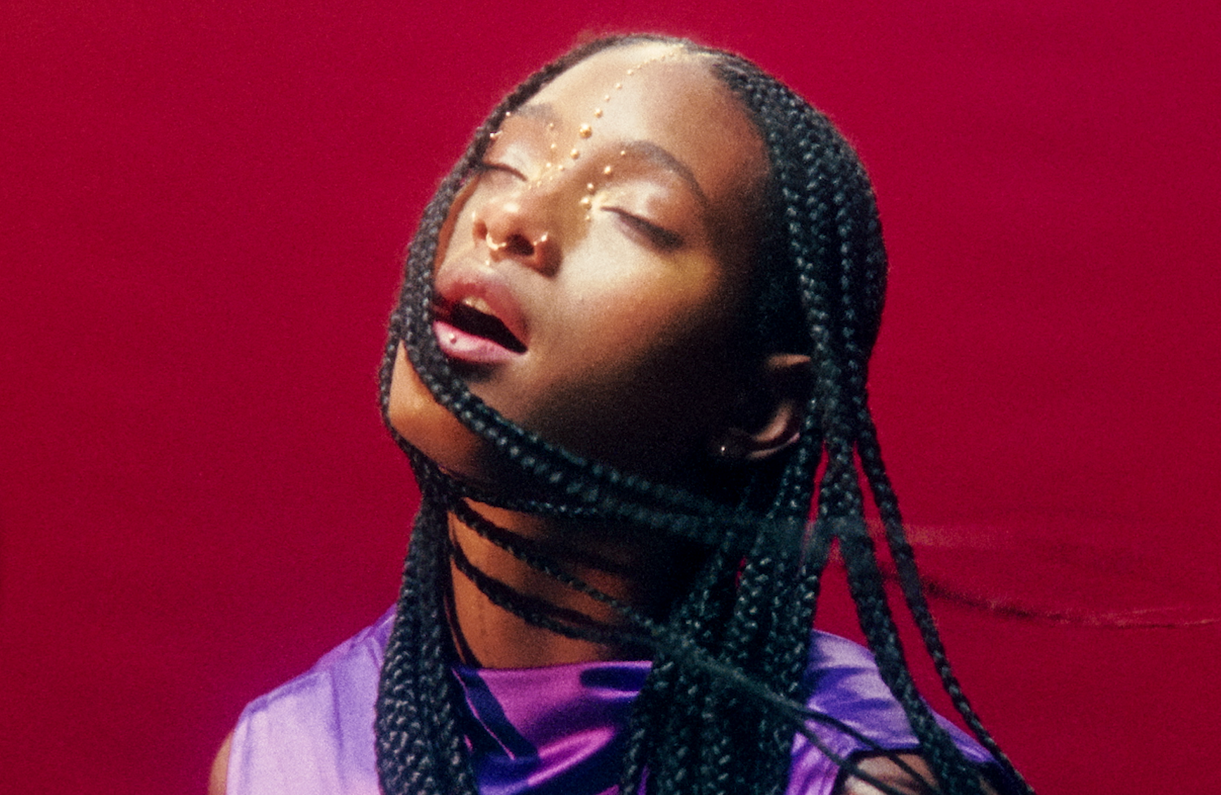 A woman with braided hair and gold face accents, with eyes closed, against a red background.