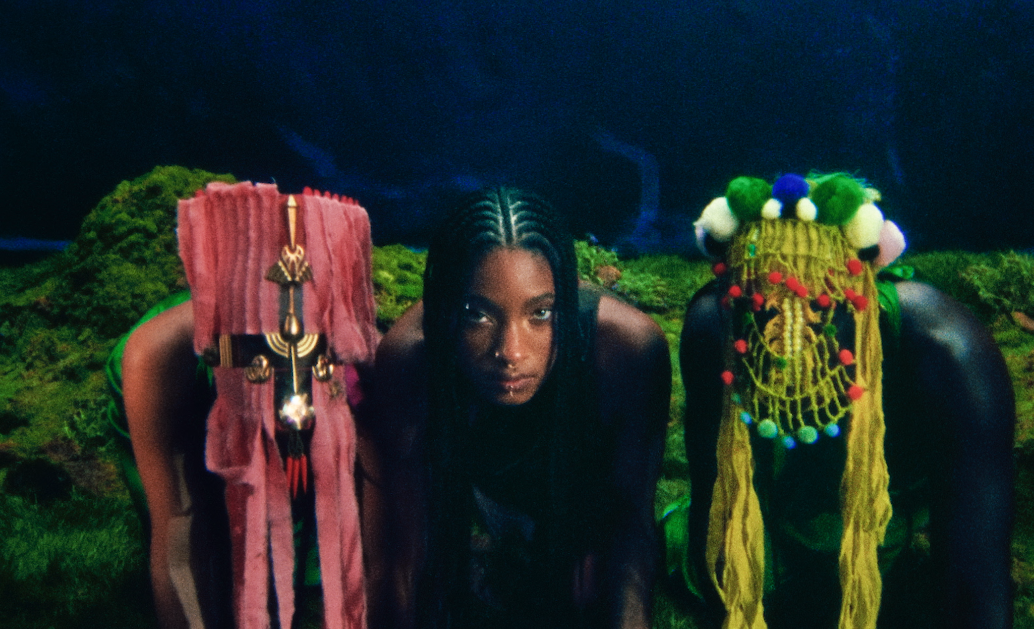 A person with braided hair in a tribal-inspired outfit with decorative accessories, accompanied by two figures with colorful, elaborate headpieces, set against a dark background.