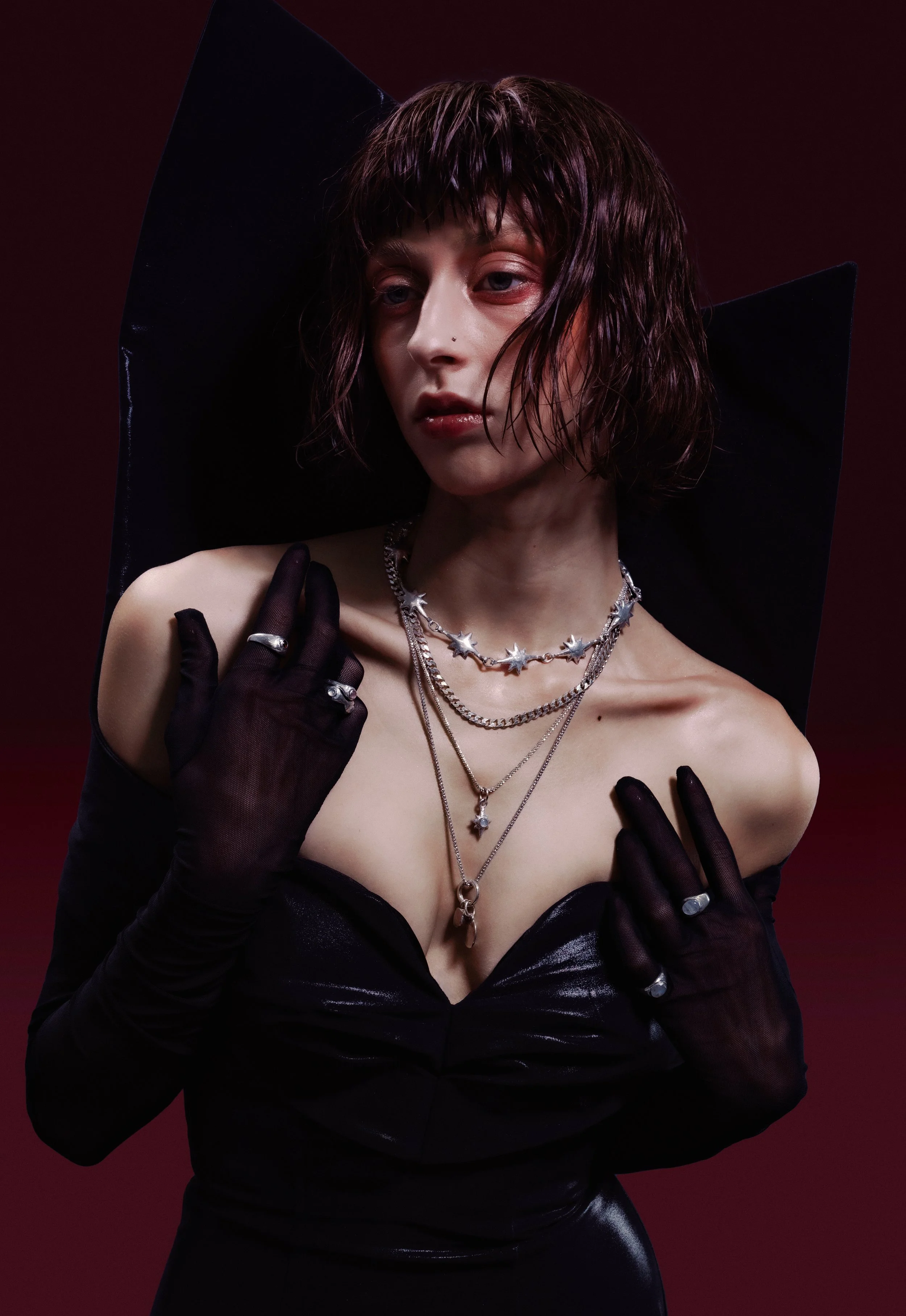 A person with short, wet-looking hair, wearing black gloves and multiple silver jewelry, including necklaces and rings, poses against a dark red background. They are dressed in a black, off-the-shoulder, shiny top with a deep neckline.