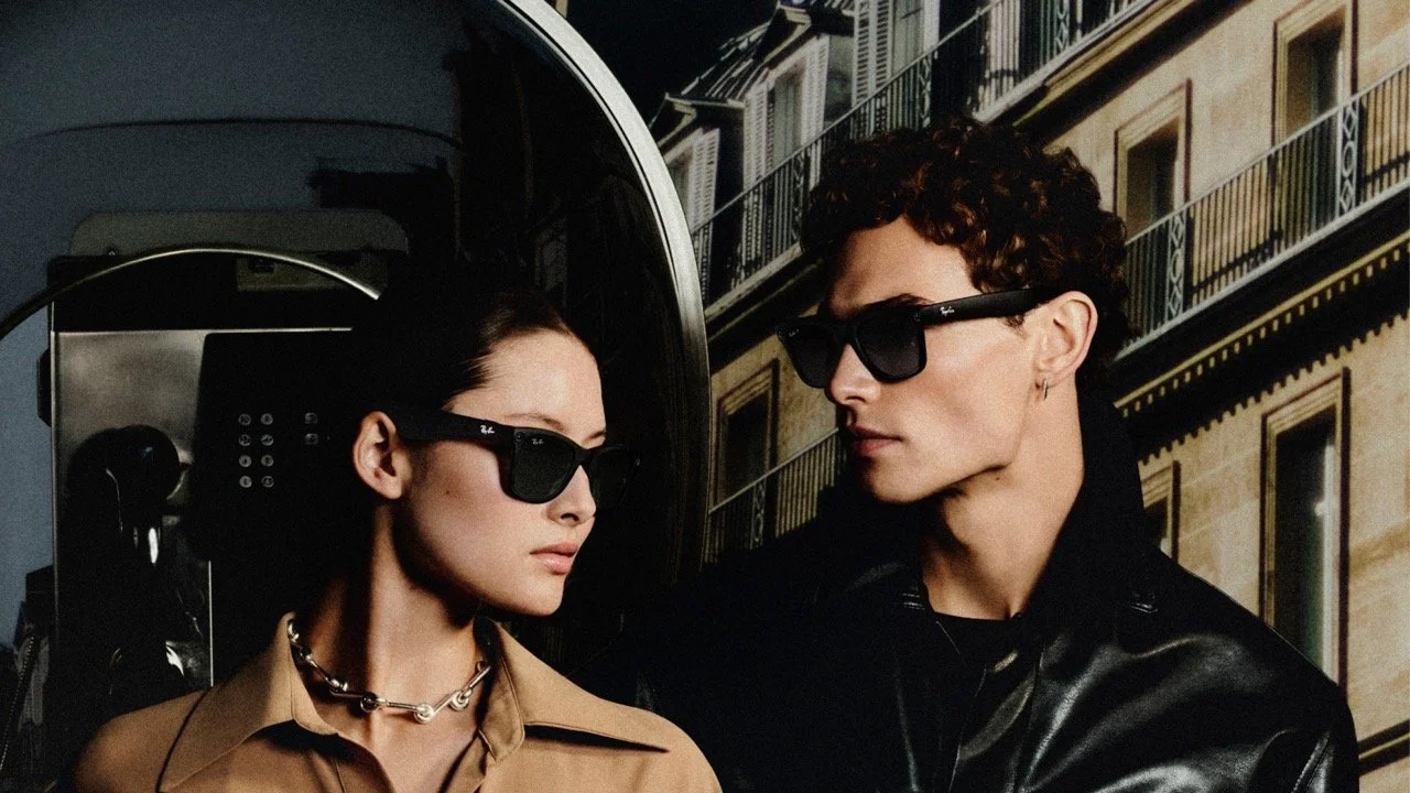 Two models with dark sunglasses stand in front of a building with balconies, one woman with dark hair in a tan shirt and a woman with curly hair in a black leather jacket.