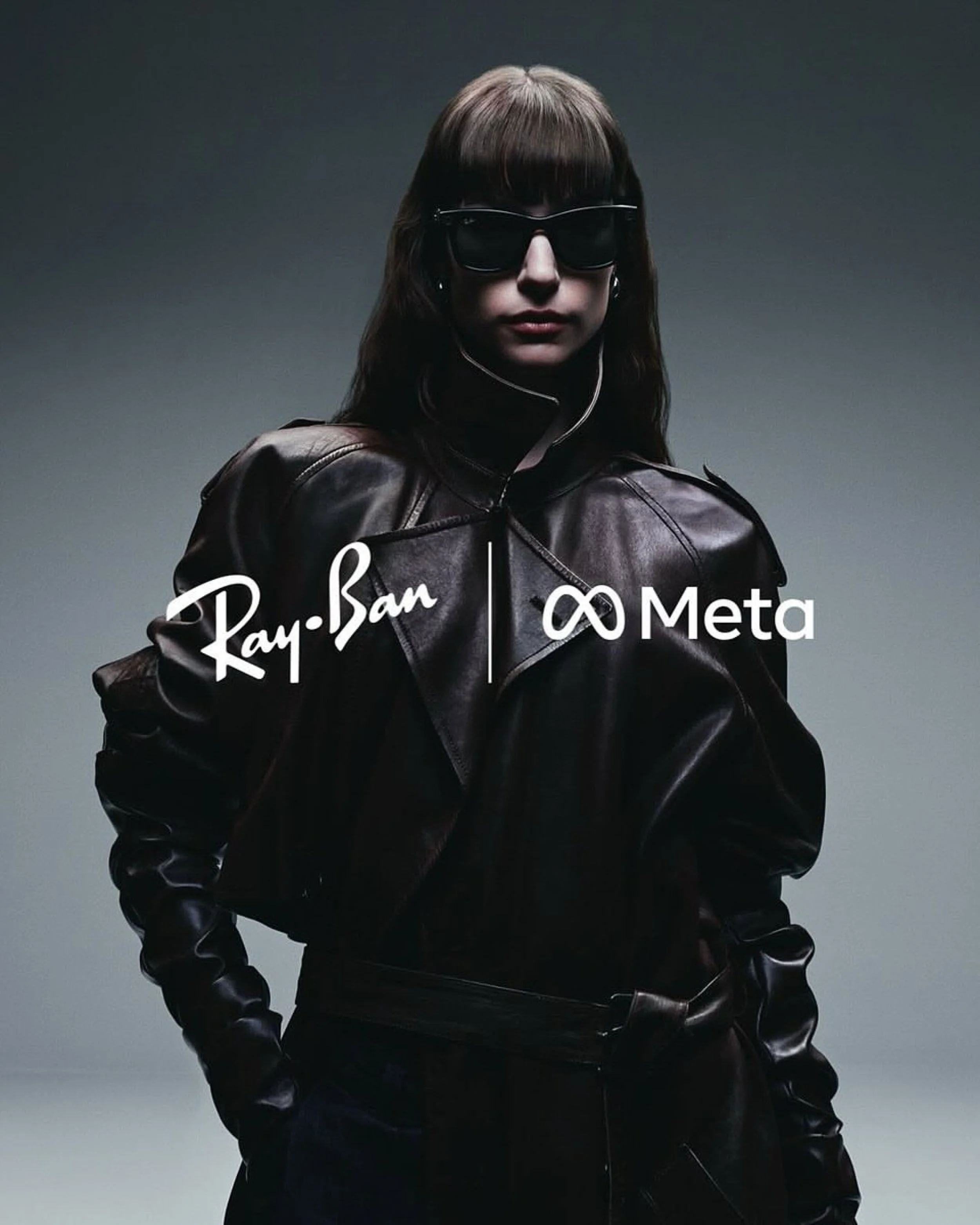A woman with dark brown hair wearing large black sunglasses, a dark leather jacket with a zippered front, and her hands in her pockets, standing against a plain gray background.