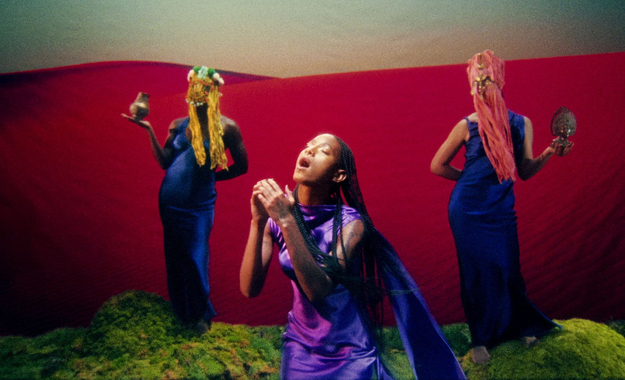 A woman with long braided hair, wearing a purple and black dress, kneeling on the ground, with two women in blue dresses holding ornate masks behind her, all standing on a grassy surface against a red and green background.