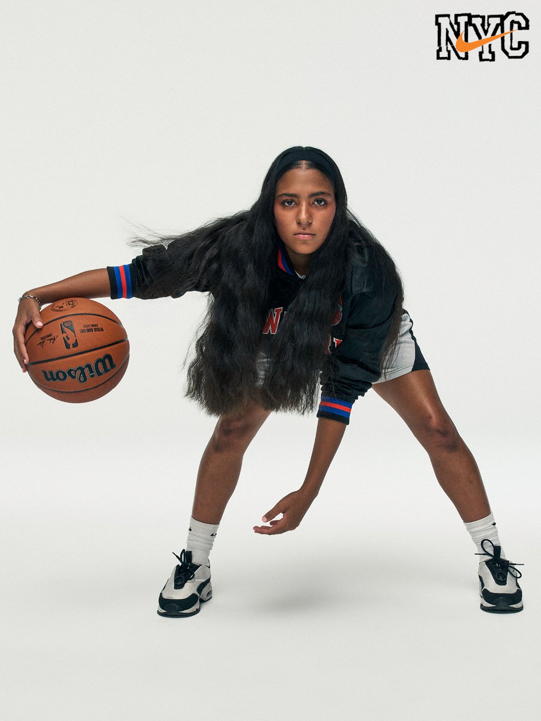 Female basketball player in black New York Knicks jacket, white shorts, and sneakers, holding a basketball with a white background.