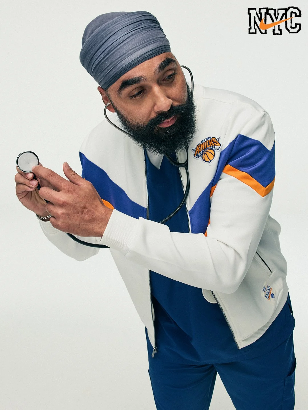 Man with a beard wearing a gray turban and a New York Knicks jacket, holding a stethoscope against his chest.