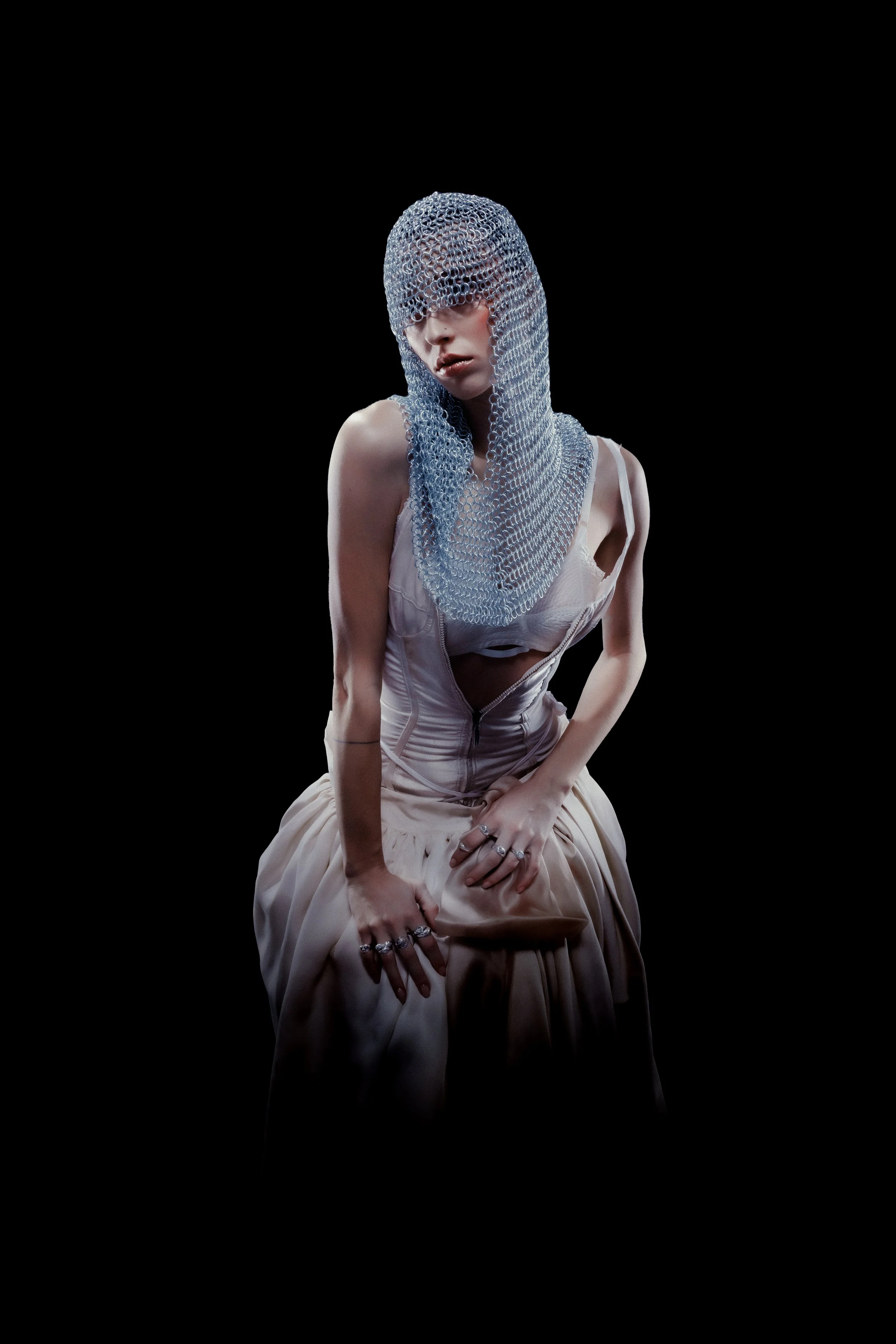 A person wearing a chainmail hood and a light-colored dress, posing against a black background.