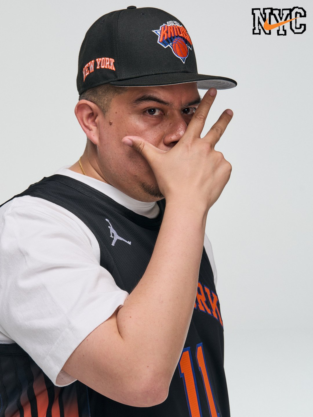 Young man wearing a New York Knicks cap and basketball jersey making a hand gesture near his face.