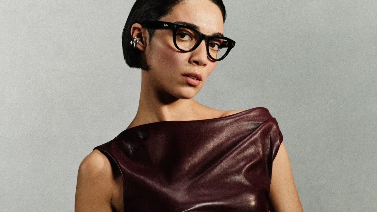 A woman with short dark hair, wearing glasses and earrings, dressed in a shiny maroon sleeveless top, poses against a gray background.
