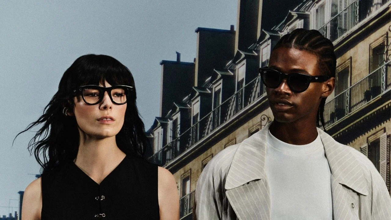 Two fashionable models wearing sunglasses and stylish clothing standing on a city street with classic European-style buildings in the background.