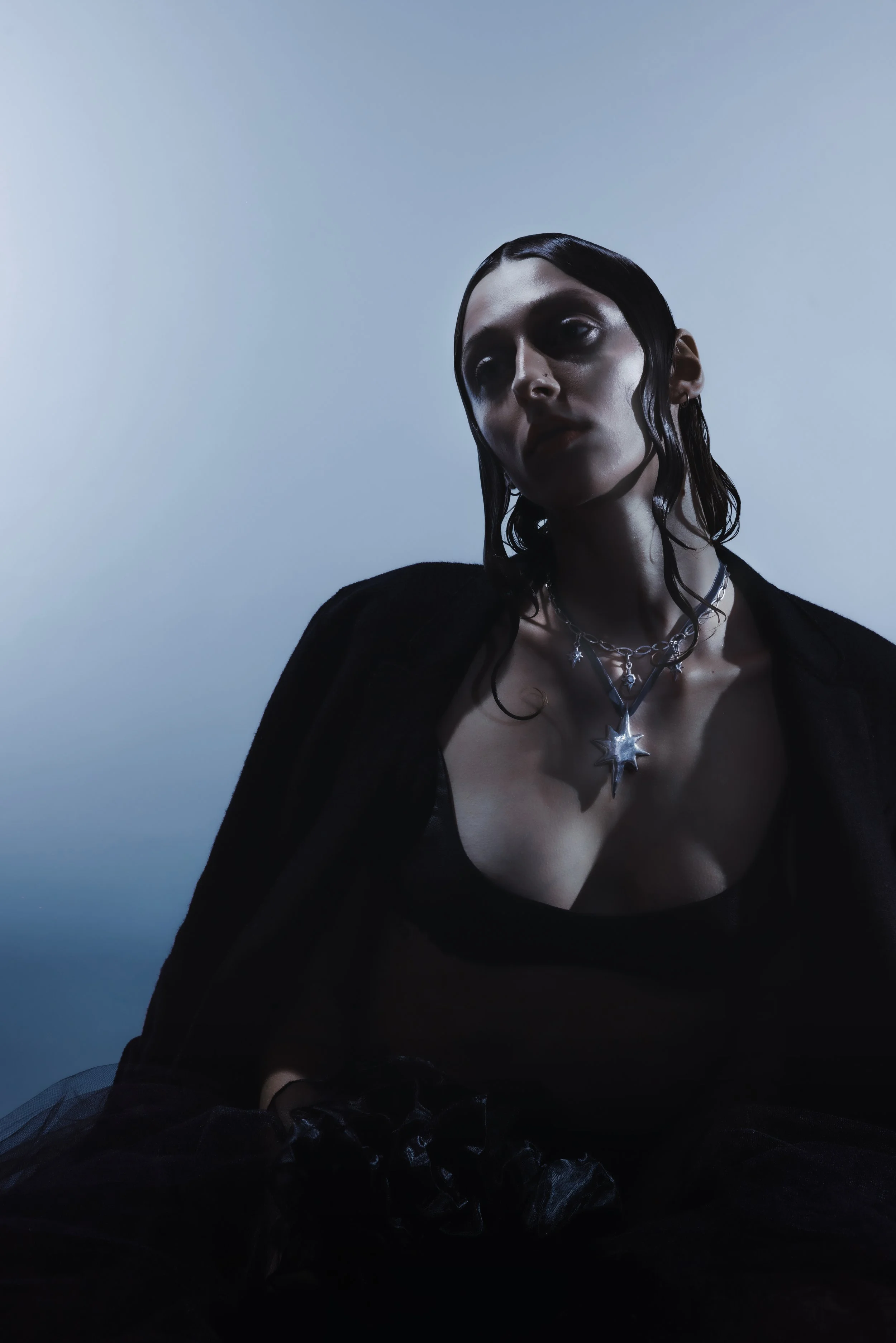 A woman with wet dark hair wearing multiple silver necklaces, including a star-shaped pendant, and a black top against a gradient blue background.