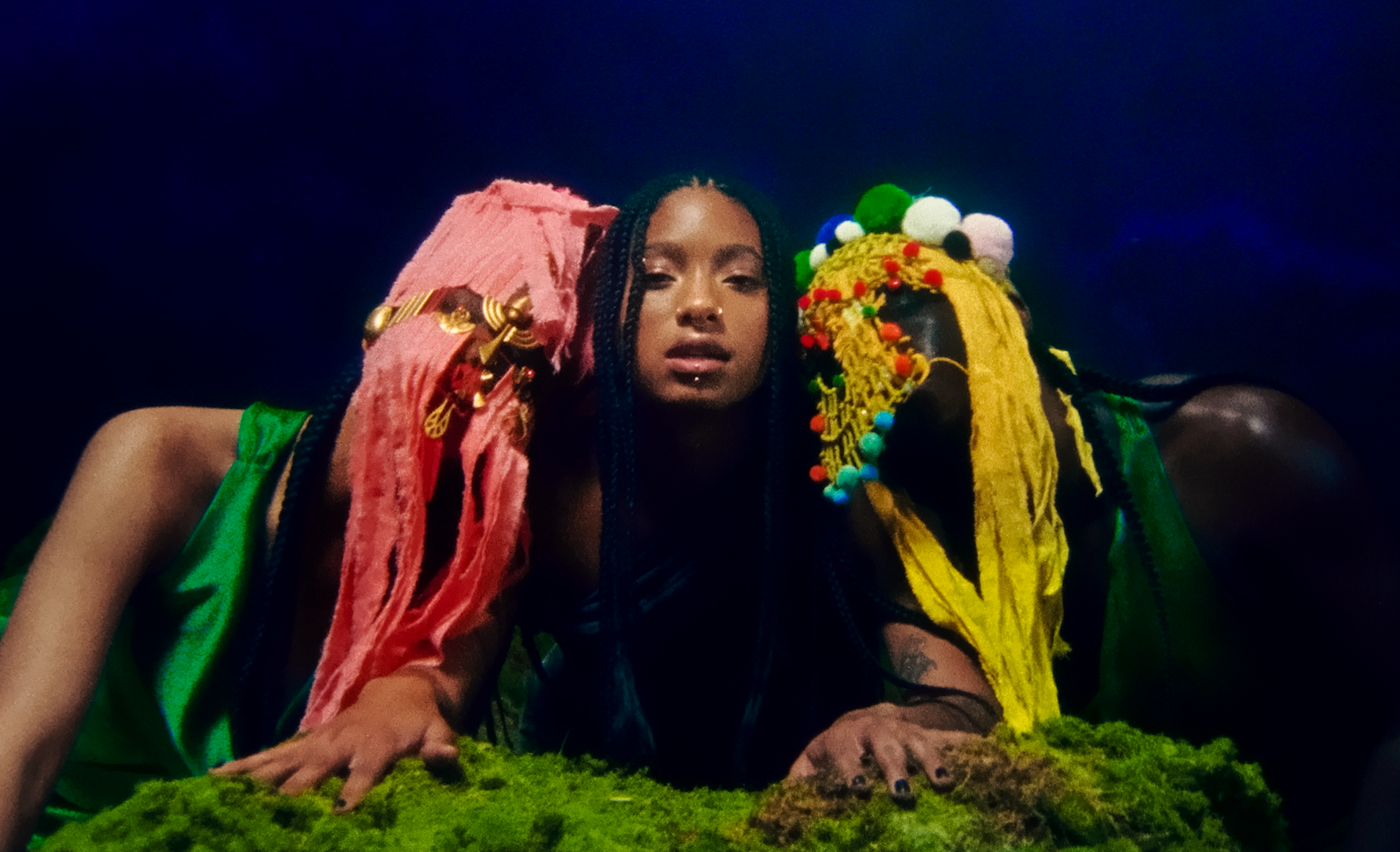 A woman with braided hair wearing a vibrant, eclectic outfit with colorful textures and accessories, standing against a dark background.