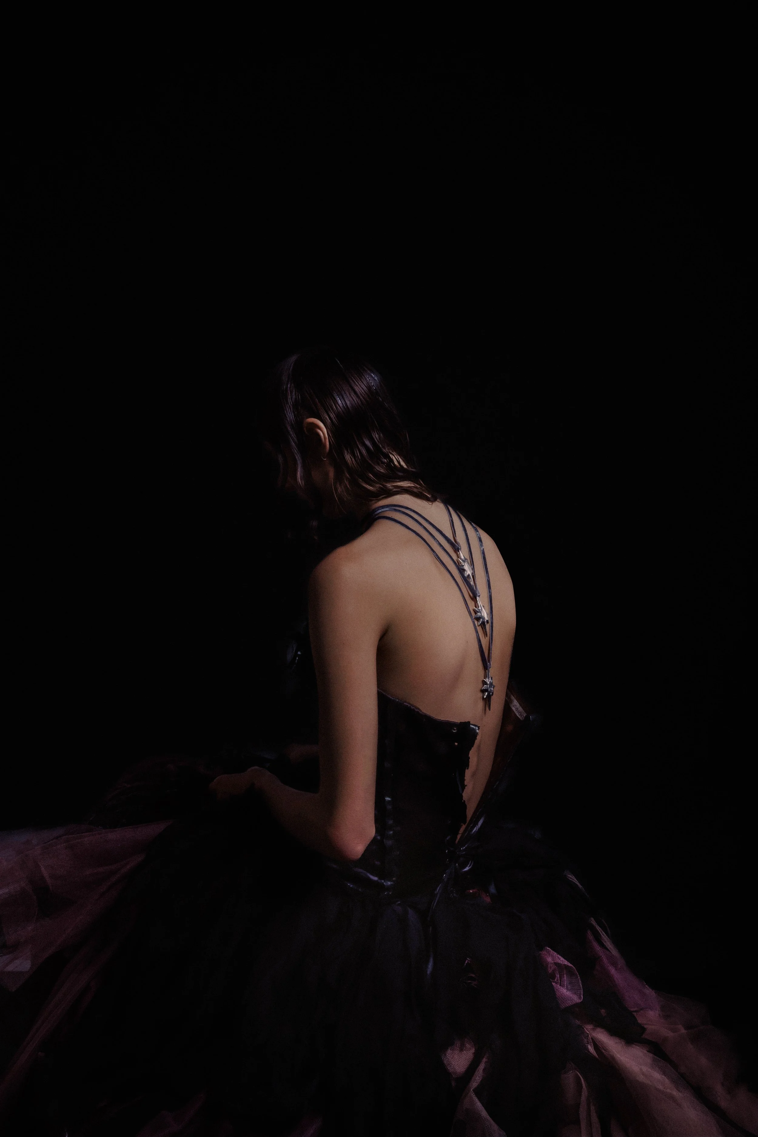 A woman with purple hair is sitting on a dark, shiny material, wearing a black dress with straps and a long necklace with star-shaped charms. The background is completely black, emphasizing her profile and attire.