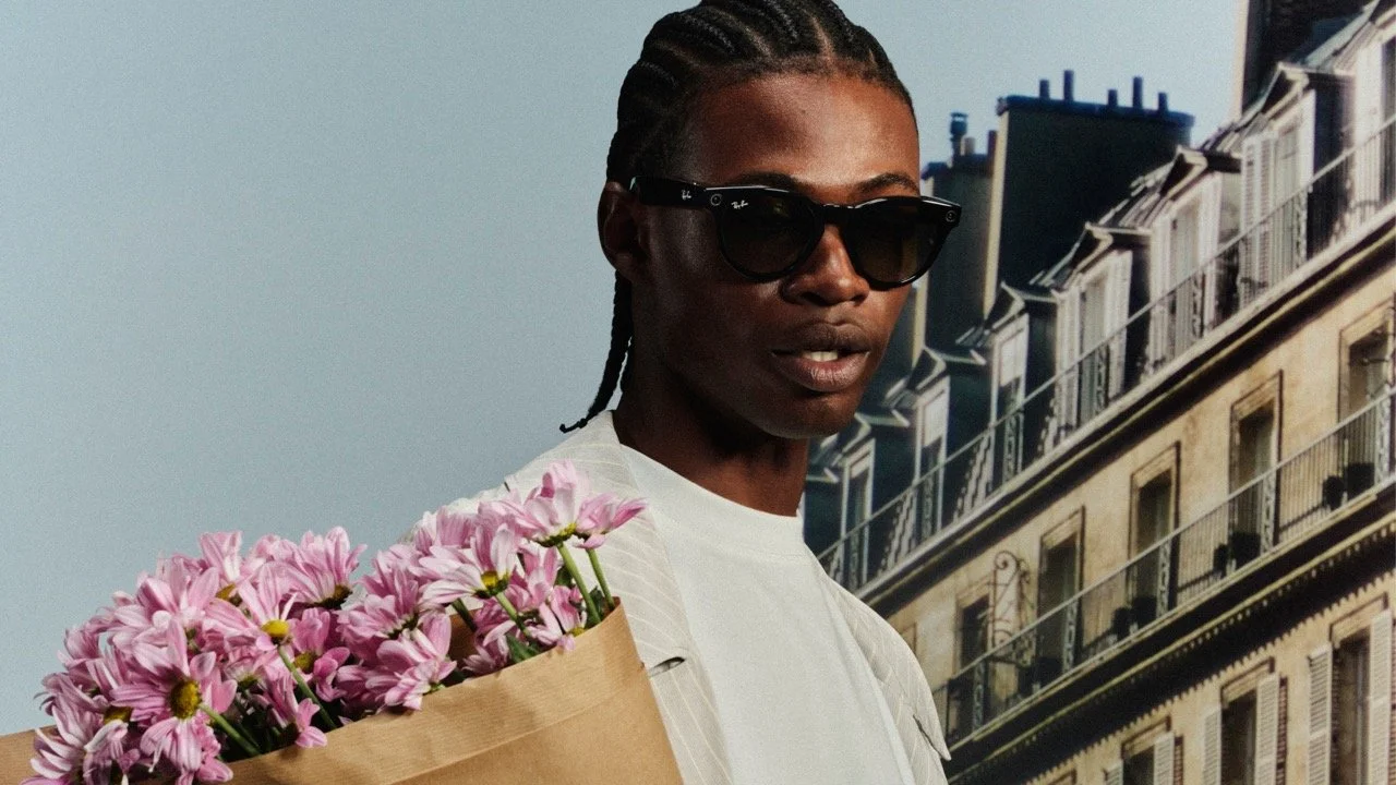 A person with dark skin and braided hair wearing sunglasses holds a bouquet of pink flowers against a background of apartment buildings and a blue sky.