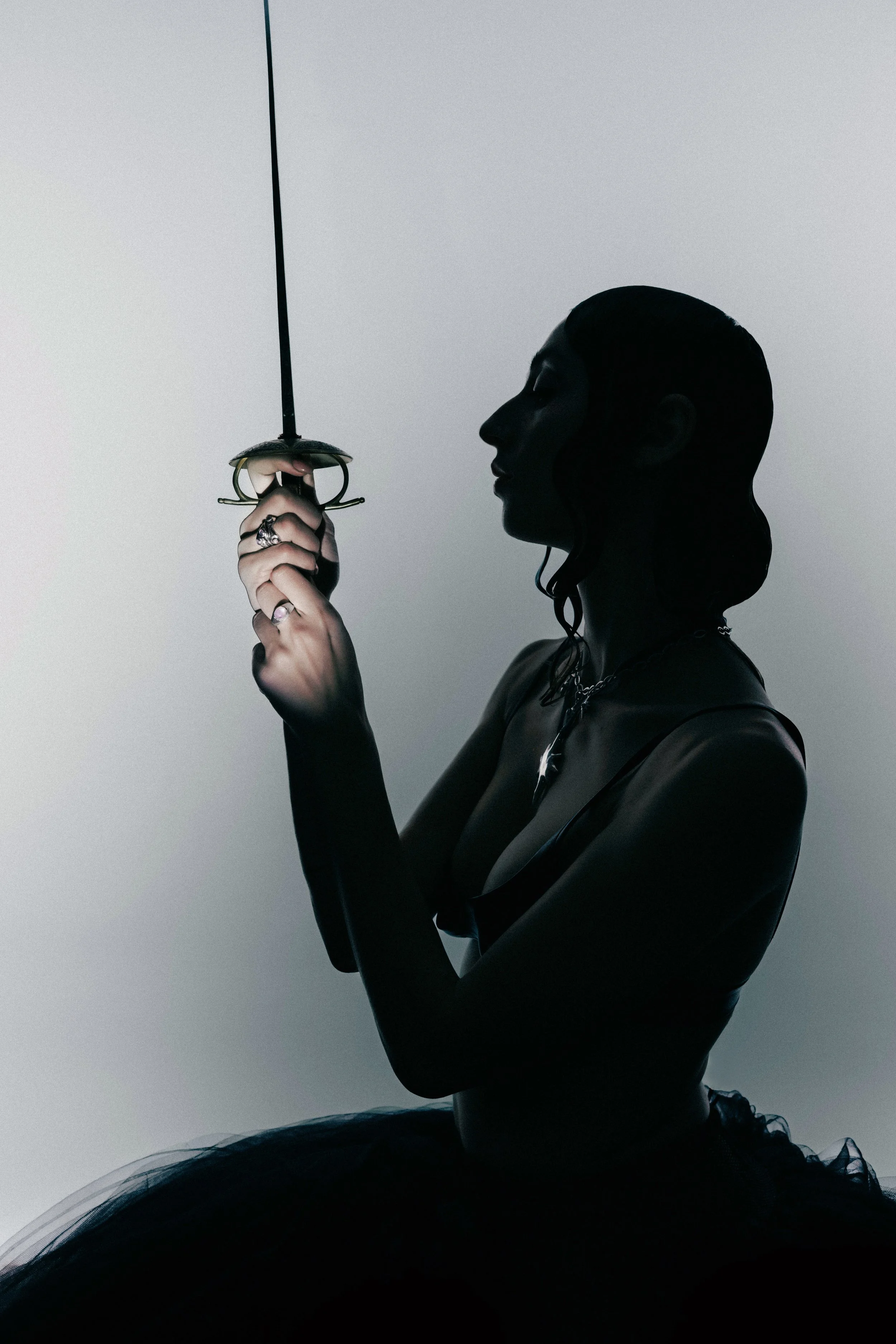 A silhouette of a woman in a black ballet tutu holding a fencing sword, with her eyes closed against a plain white background.