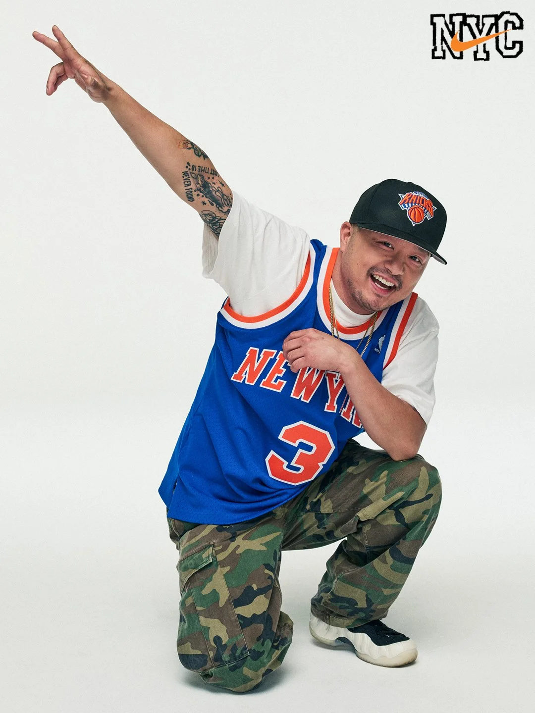 A man wearing a New York Knicks basketball jersey, camouflage pants, and a black Knicks cap, crouching on one knee with a playful pose, smiling against a plain white background.