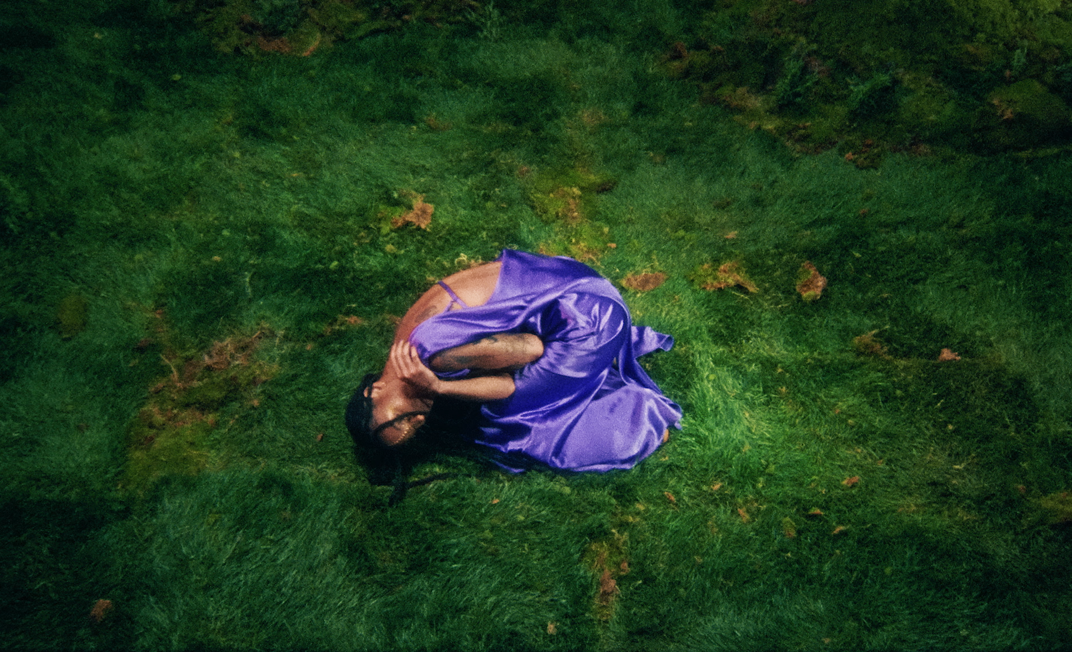 A person wearing a purple satin dress lying on green grass, covering their face with their hands.