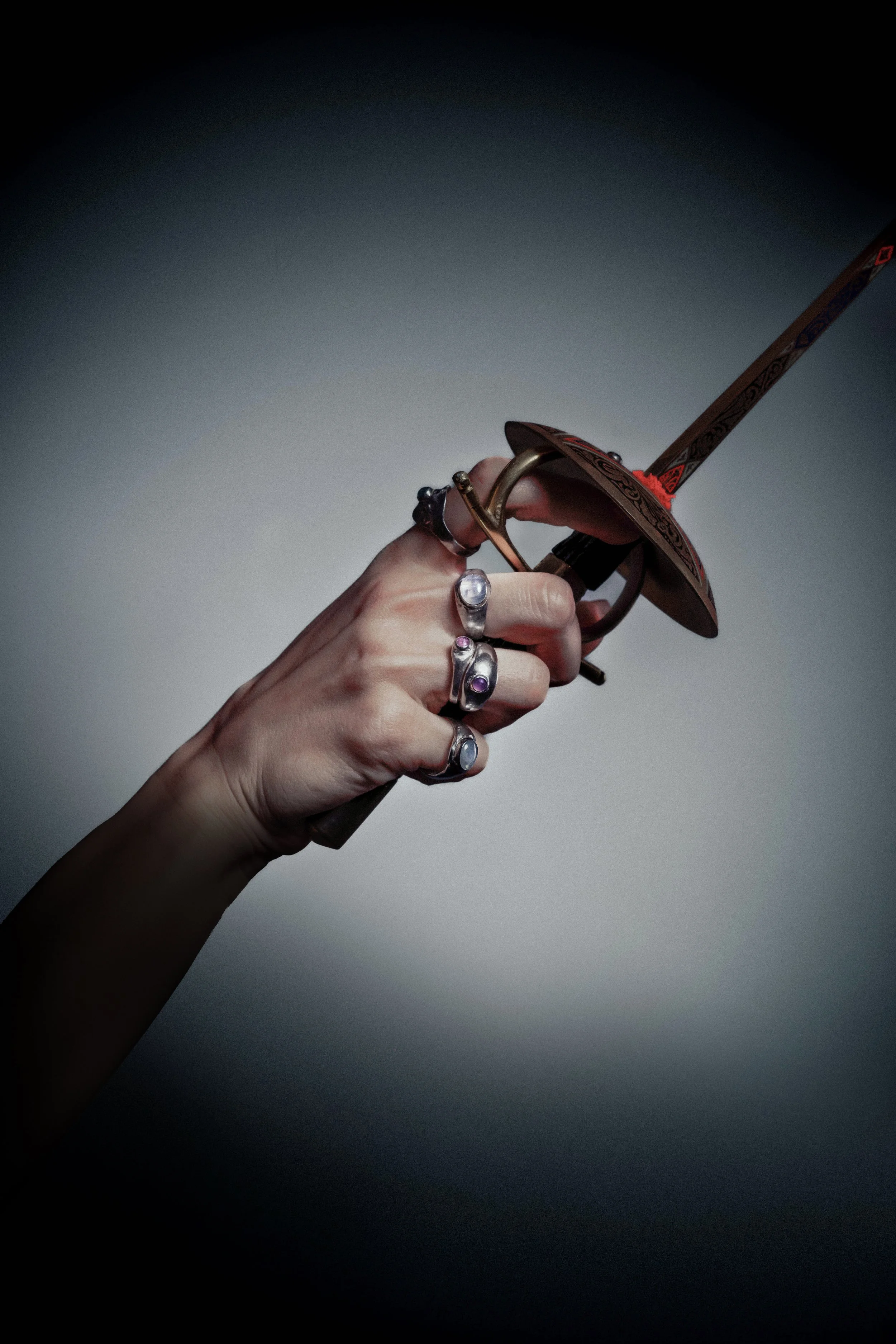 A hand holding a decorative sword with a patterned blade and ornate guard, adorned with silver rings with gemstones.