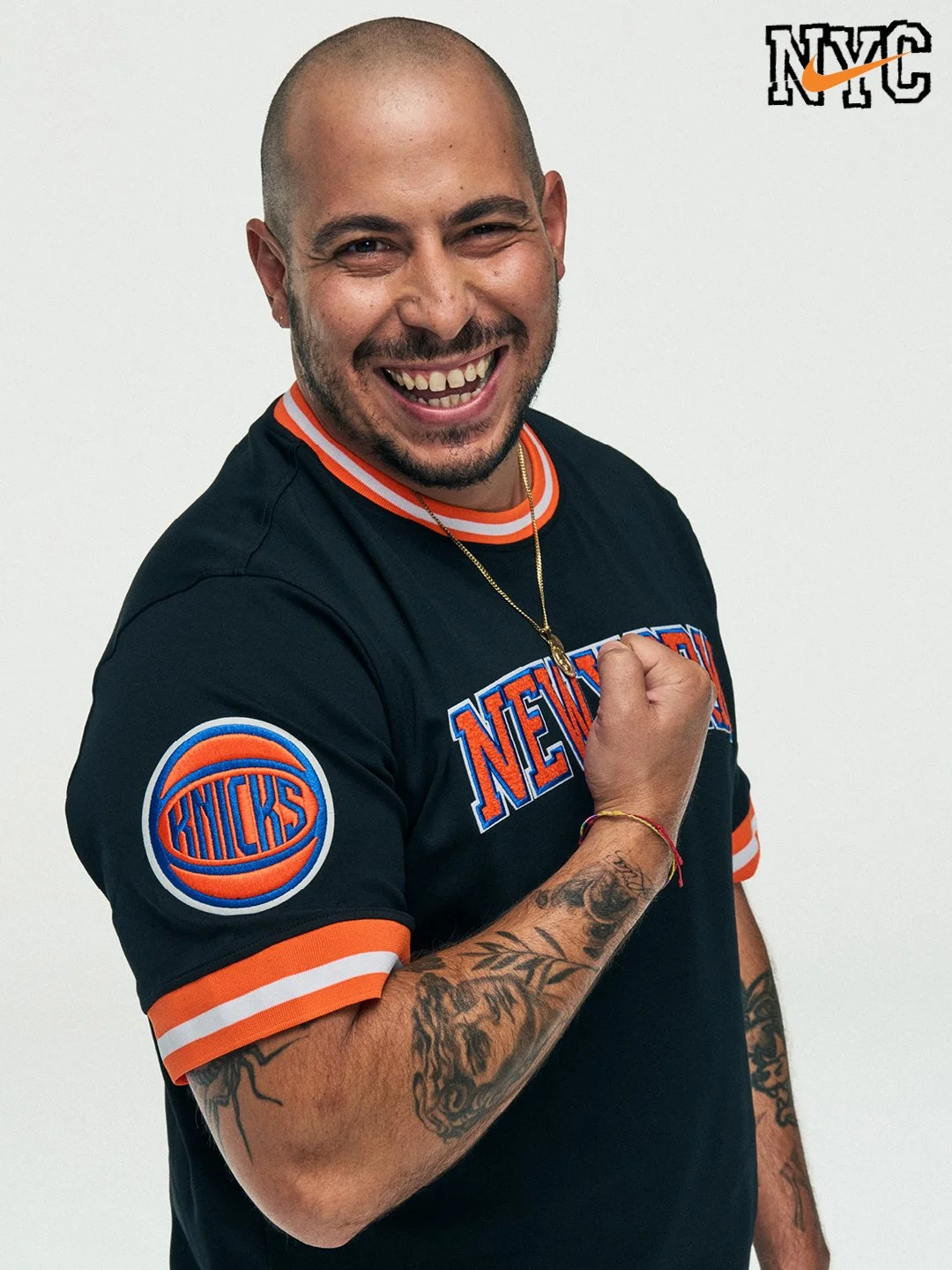 A man smiling and showing a fist in a New York Knicks sports jersey, with visible tattoos on his arm, against a plain white background, with NYC and Nike logos in the top right corner.