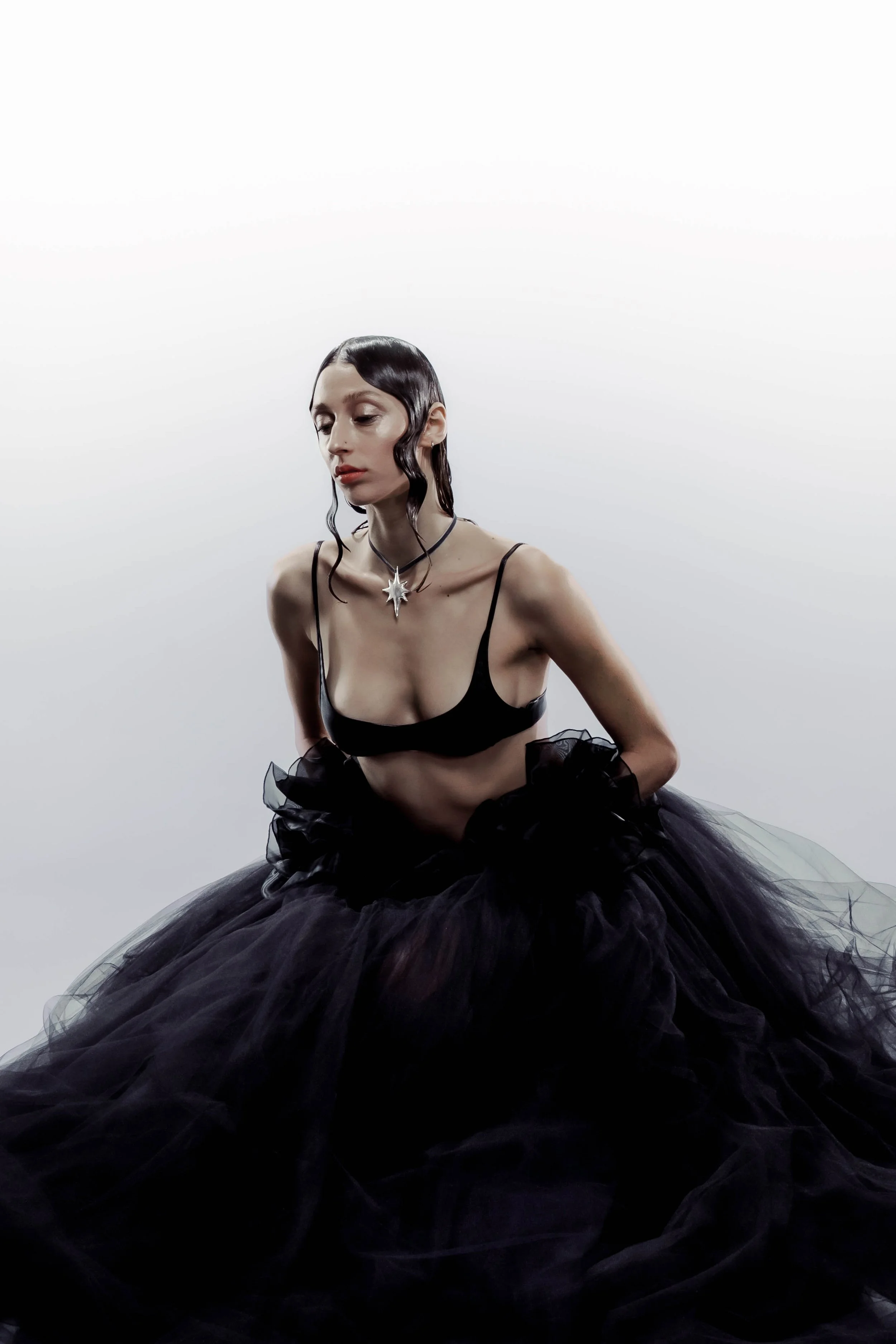 A woman with black hair styled in waves wearing a black crop top and a voluminous black tulle skirt, accessorized with a silver star-shaped necklace, sitting against a plain white background.