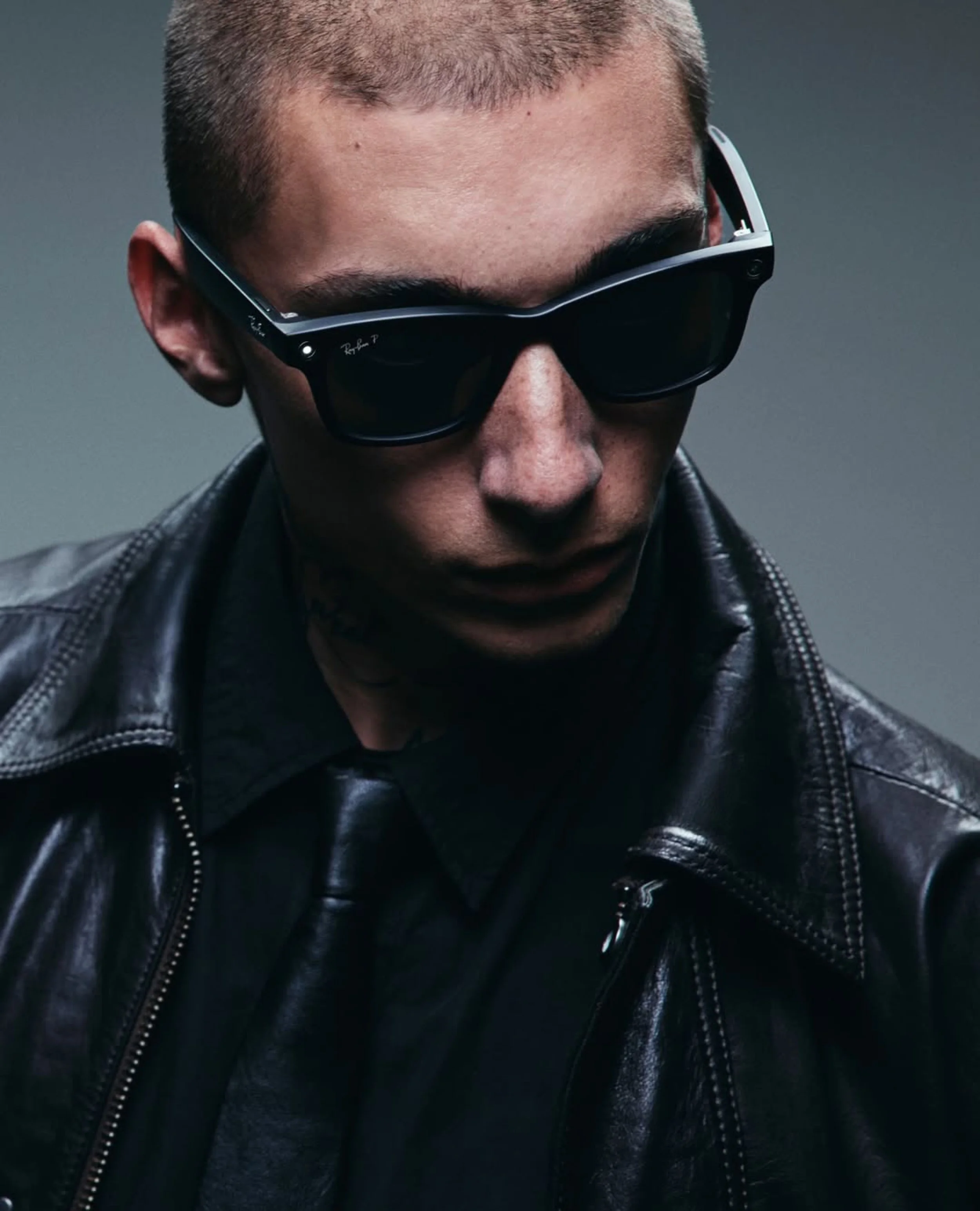Close-up of a young man wearing black sunglasses and a black leather jacket, with short hair and a serious expression.