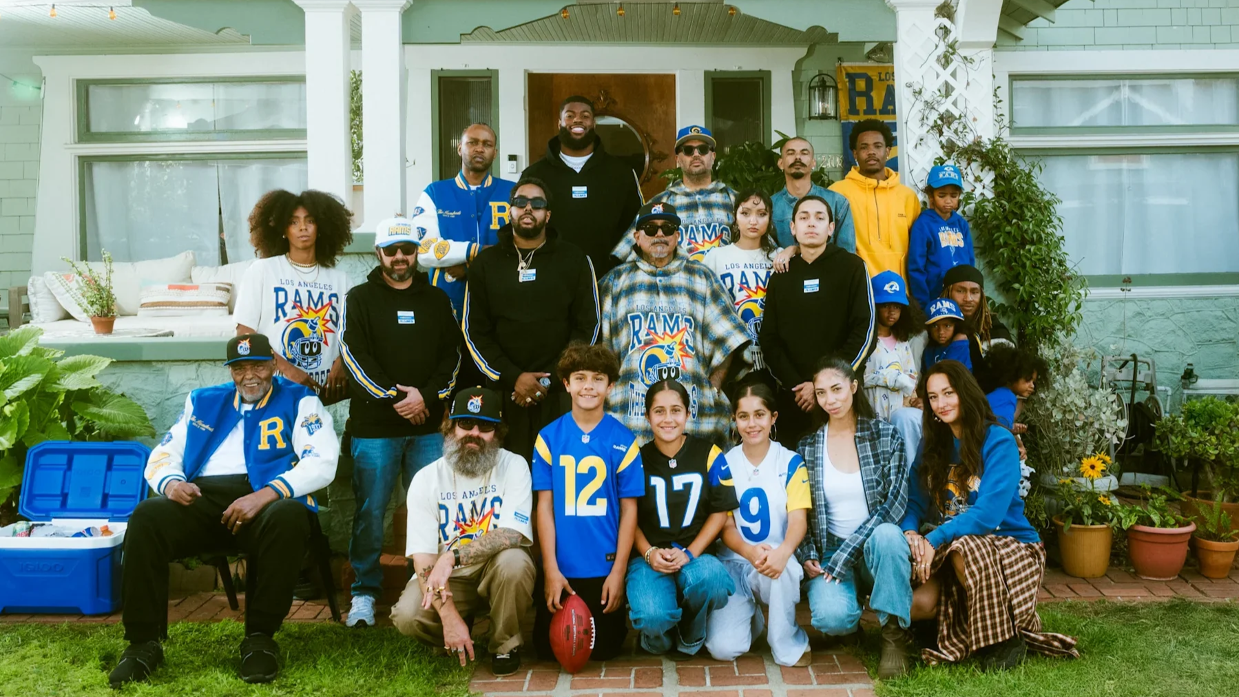 Group of people, including children and adults, gathered on the front porch of a green house, some wearing Los Angeles Rams football jerseys and apparel, posing for a photo during daytime.