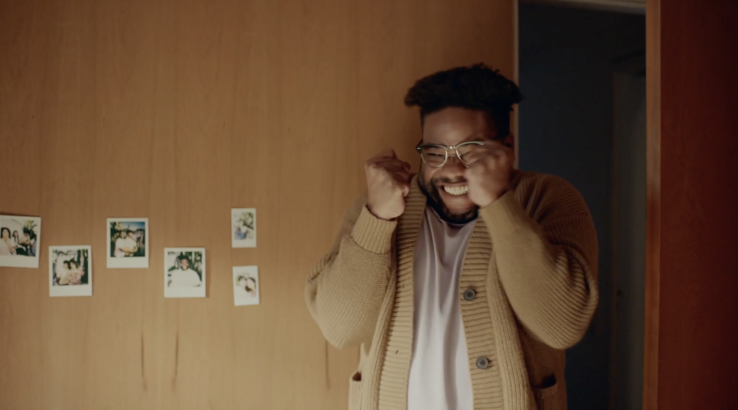 A man with glasses and a beard, wearing a beige cardigan, joyfully reacts with clenched fists, standing in front of a wall with photo prints.