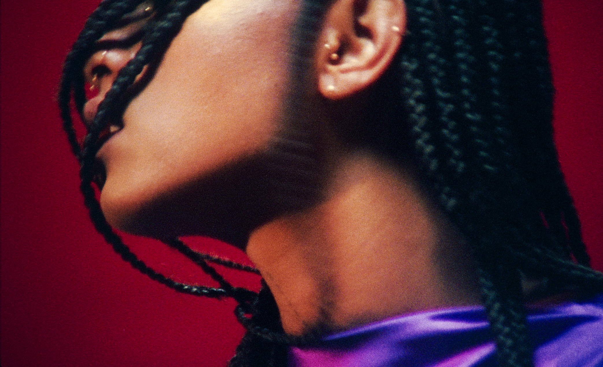 Close-up of a person's face, showing lips, ear, and braided hair against a red background.