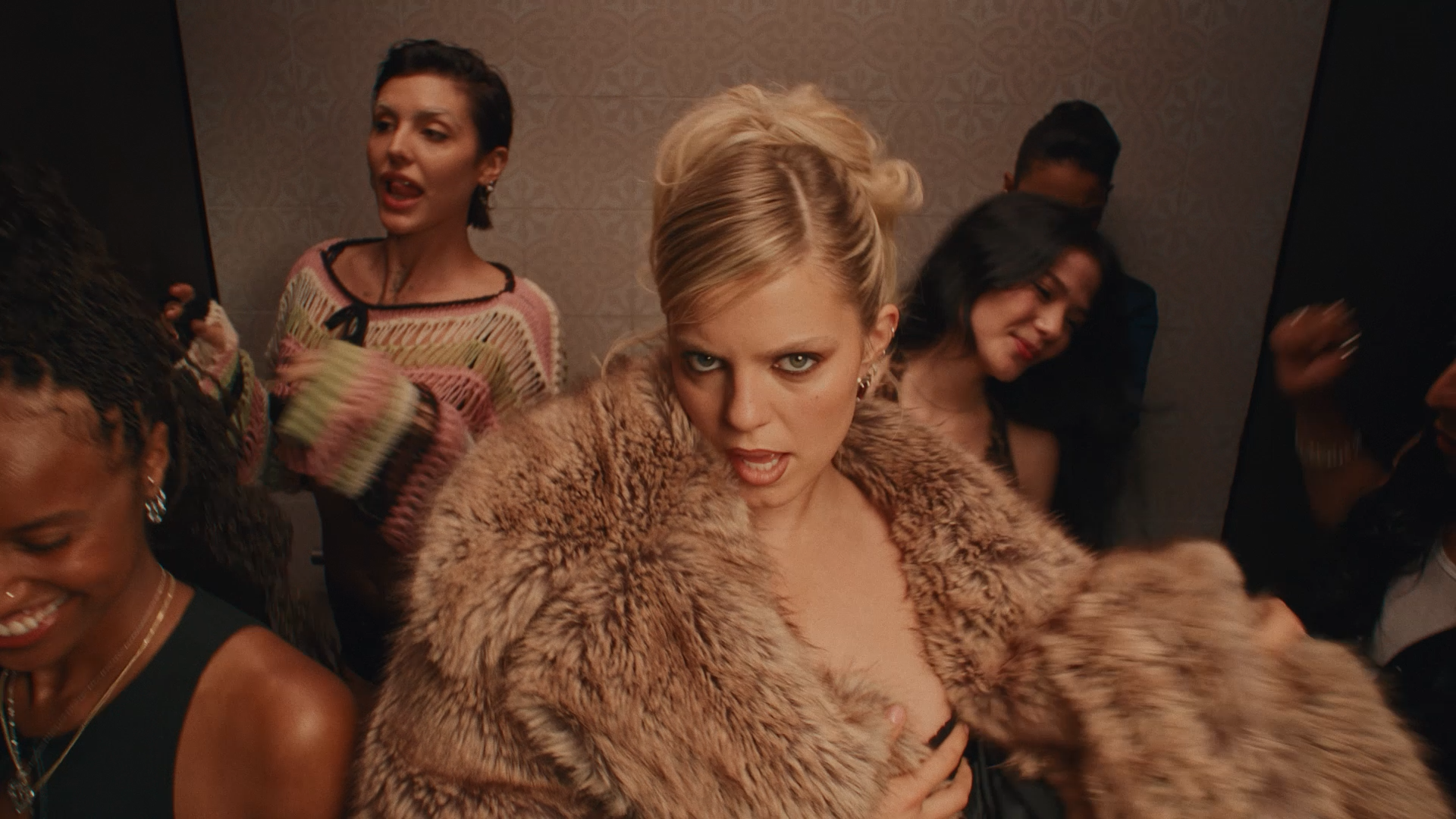 A group of women at a party, with a blonde woman in the center wearing a fur coat and making a playful, intense facial expression.