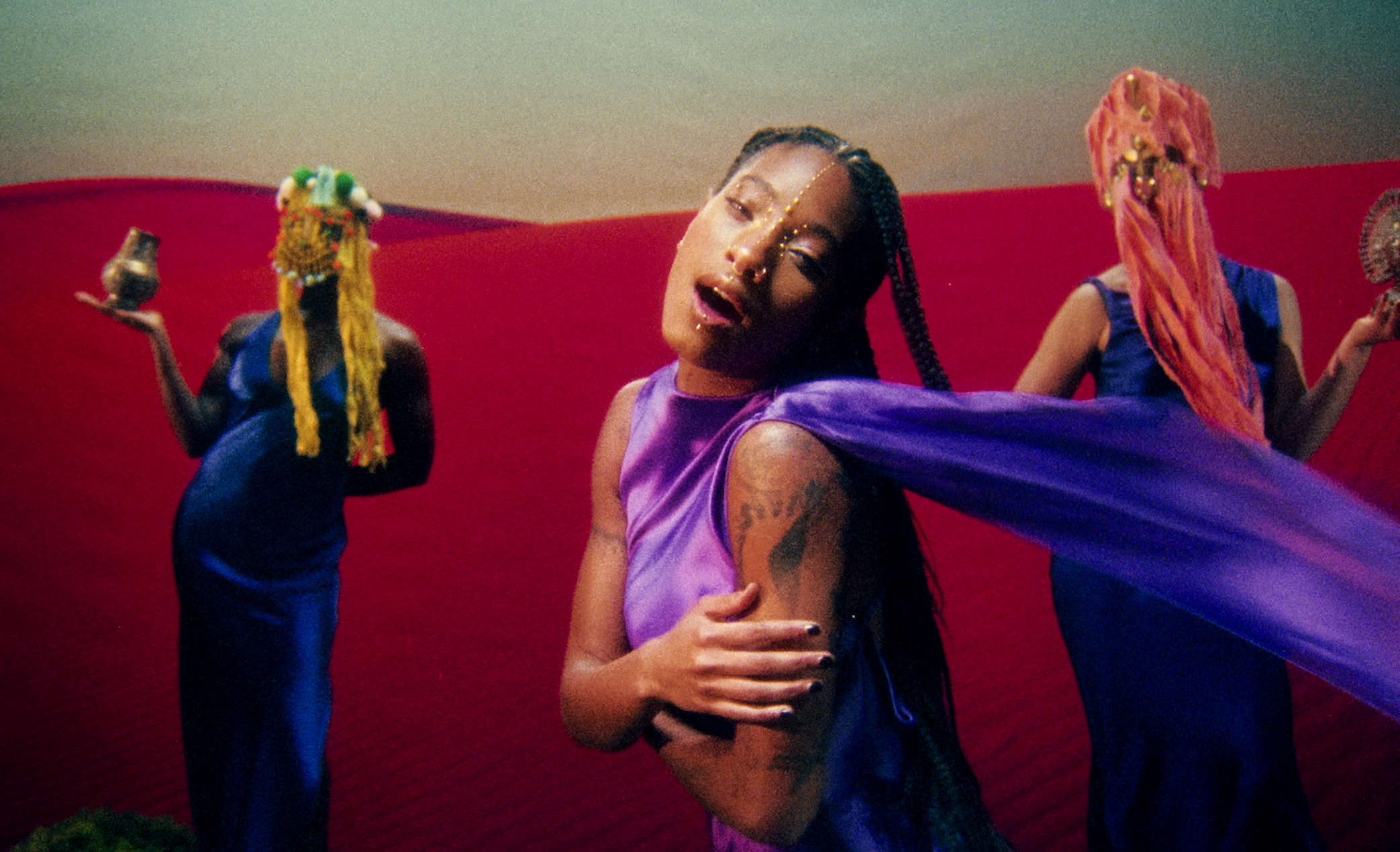 A woman with braided hair and face jewelry, wearing a purple dress, surrounded by two people with colorful head coverings holding small items, sitting against a red backdrop.