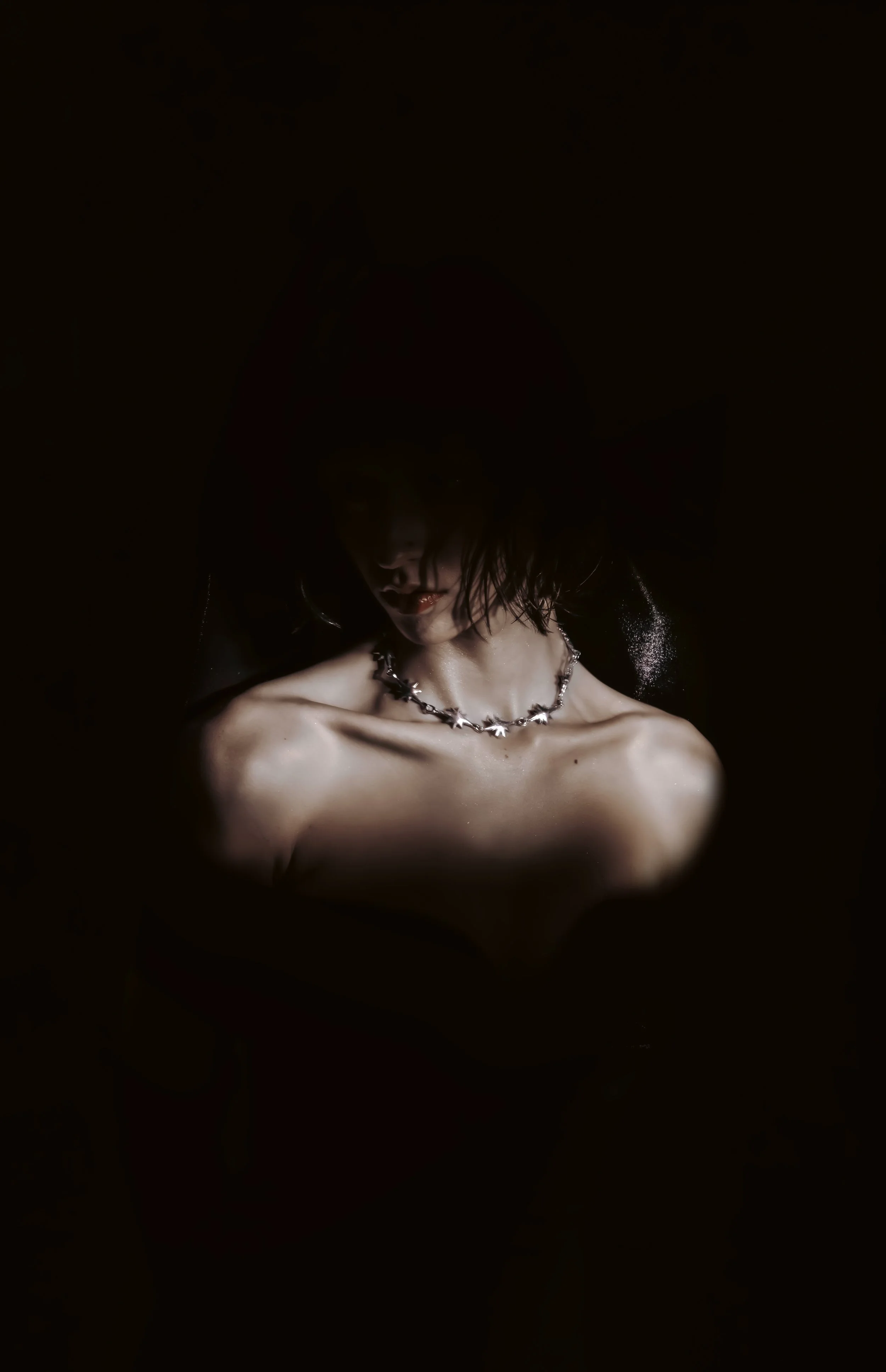 A woman with dark hair and makeup, wearing a star-shaped necklace, is topless with her shoulders exposed, against a dark background.