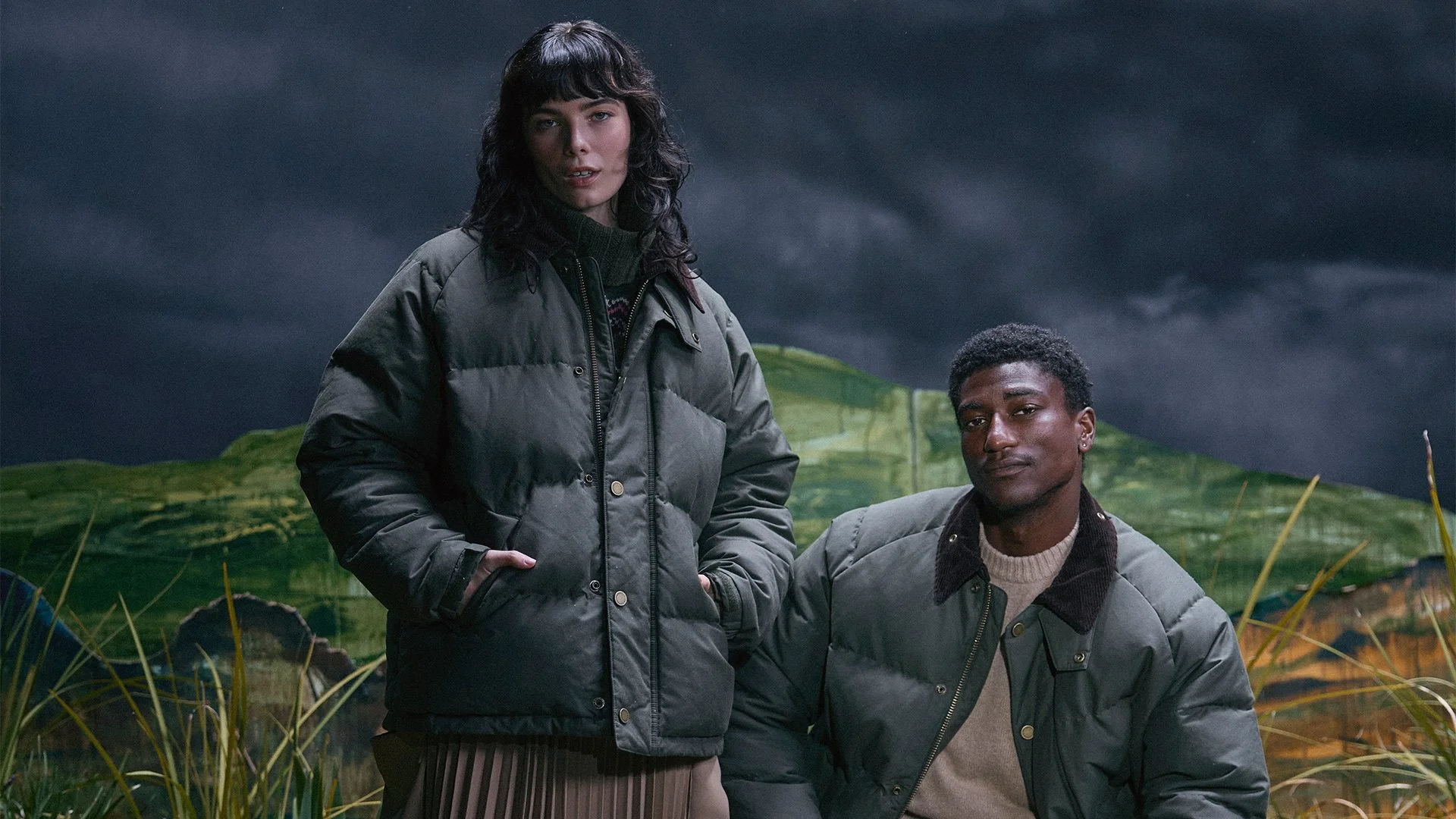 Two young adults, a woman and a man, standing outdoors in a landscape with green hills and a cloudy, stormy sky. The woman has dark hair with bangs and is wearing a gray puffer jacket and a patterned skirt. The man has short curly hair and is wearing a similar gray jacket over a cream-colored sweater. They are posing confidently amidst a moody, natural setting.