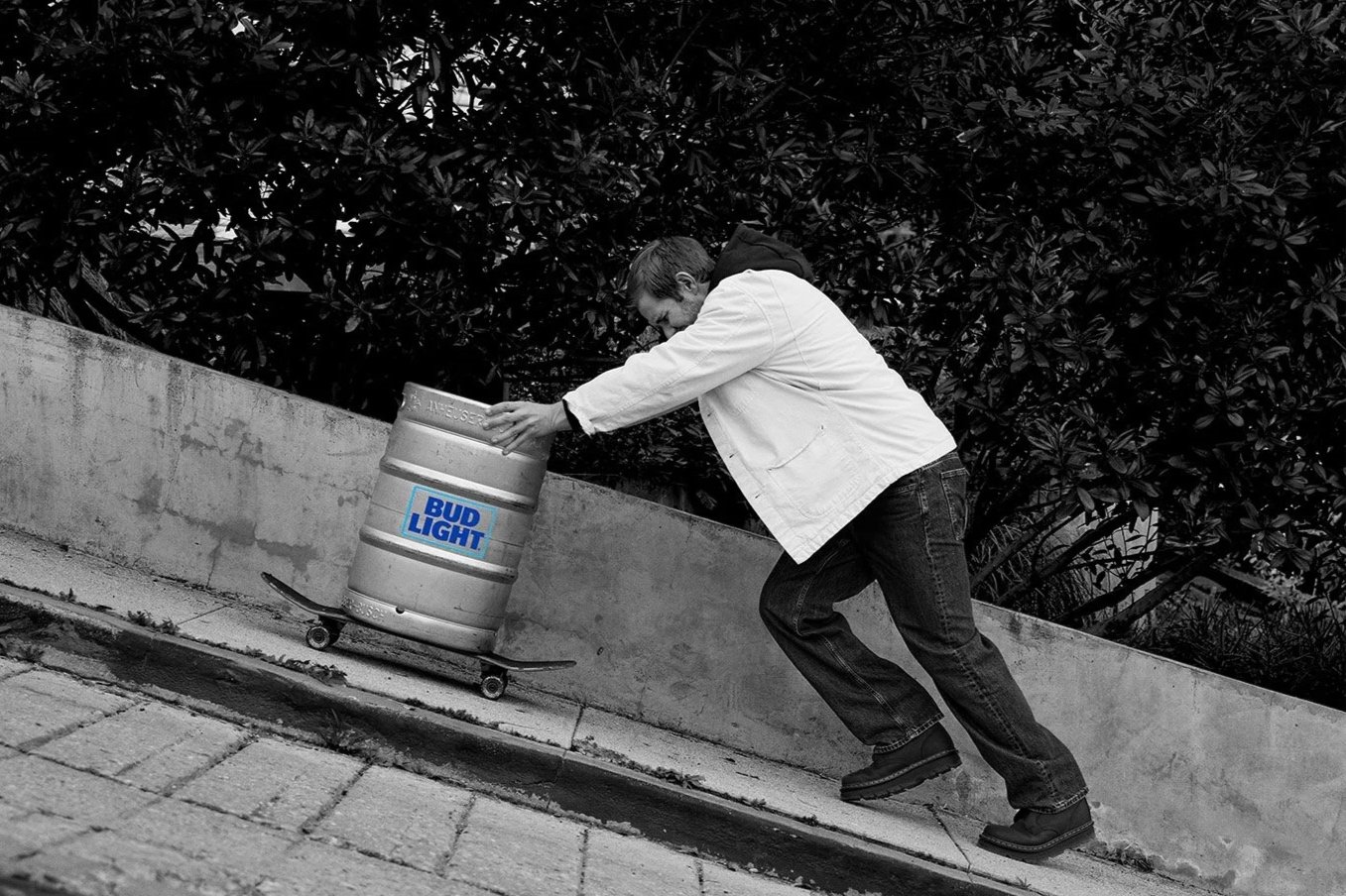A man in a white jacket and jeans rolling a skateboard up a sloped sidewalk while pushing a trash barrel labeled Bud Light.