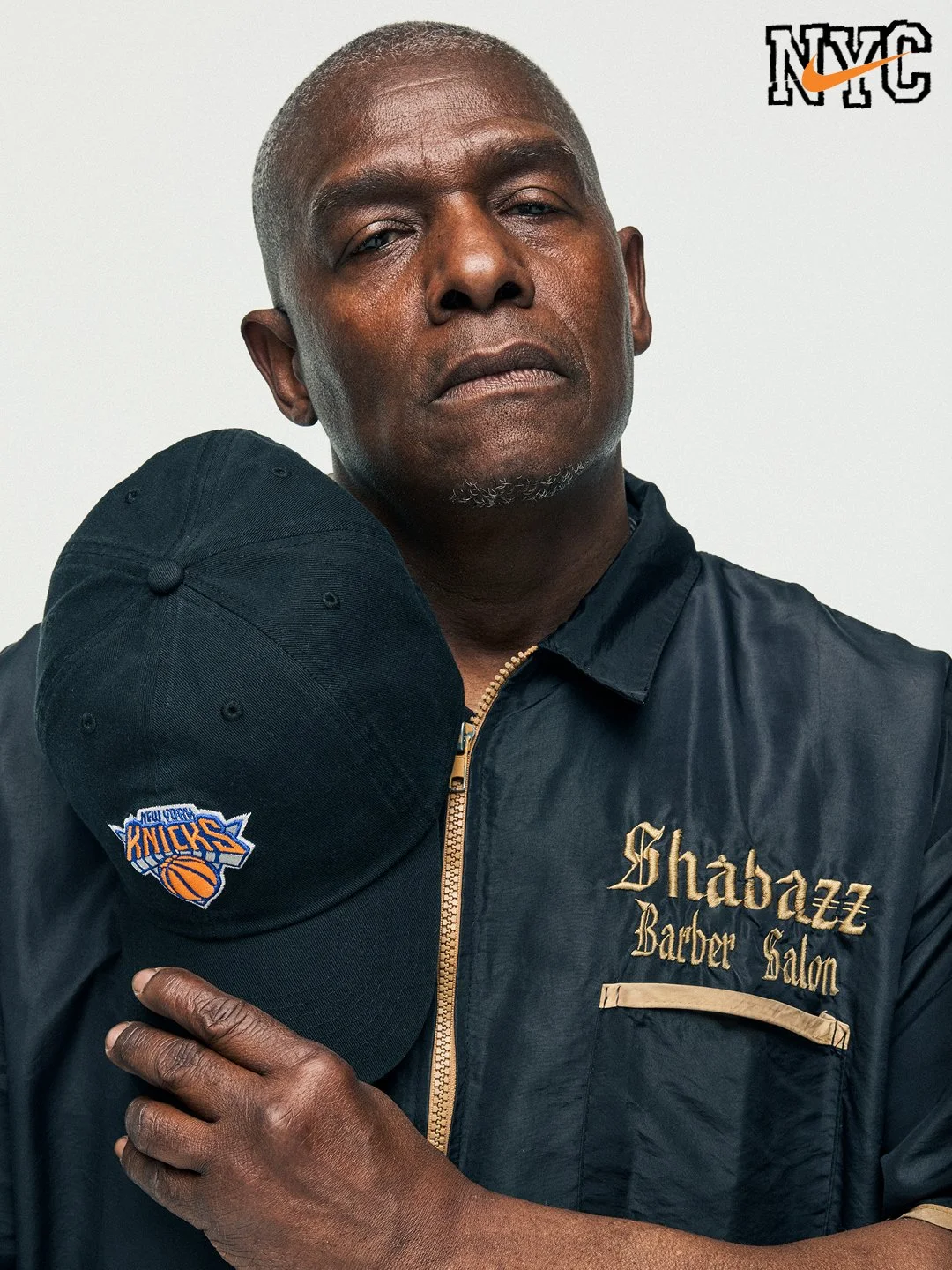 A man holding a New York Knicks cap on his shoulder, wearing a black jacket with gold embroidery that reads Shabazz Barber Salon, with a NYC logo in the top right corner.