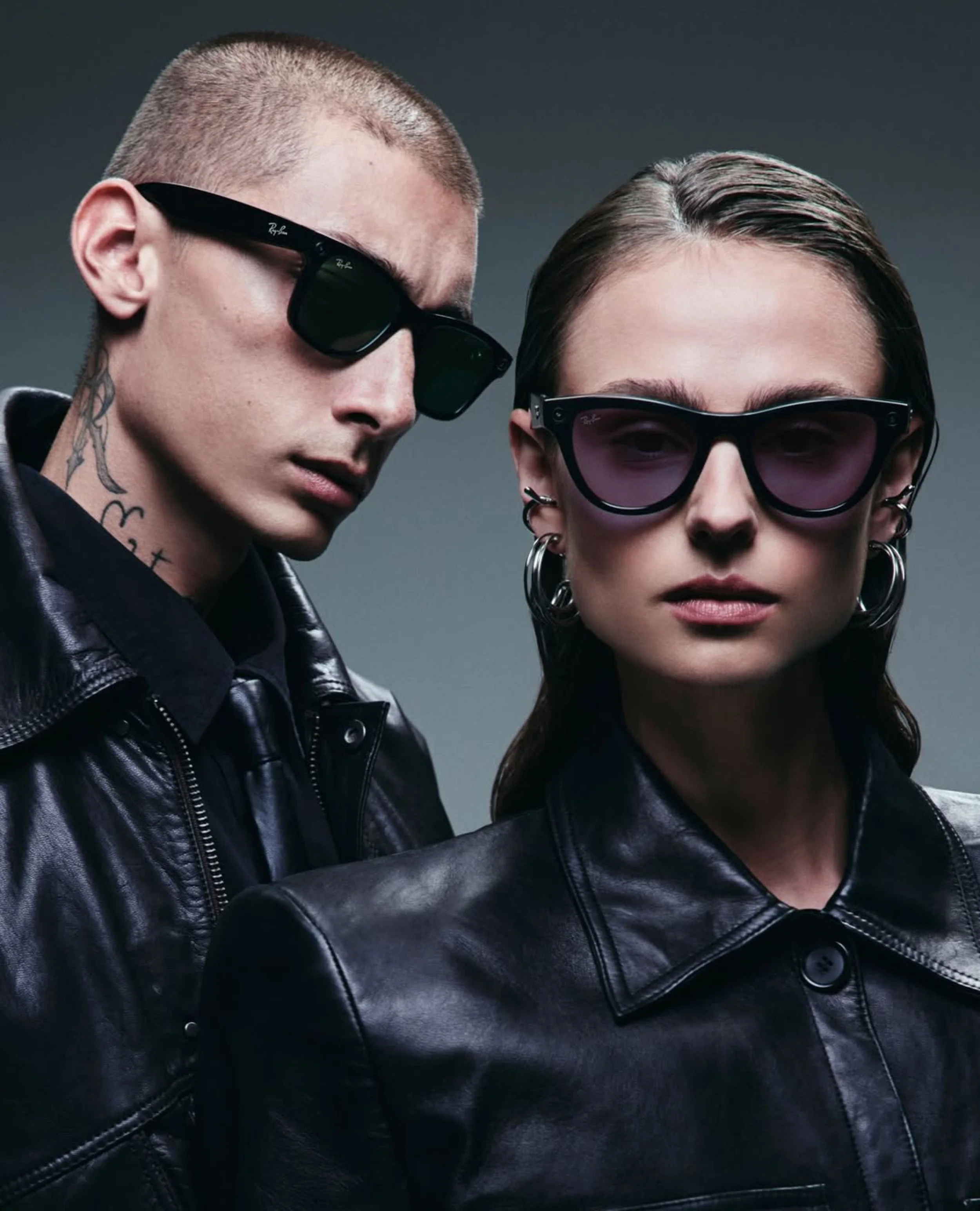 Close-up of a man and woman wearing black sunglasses and black leather jackets, with serious expressions. The man has a buzz cut, tattoos on his neck, and is wearing Ray-Ban sunglasses. The woman has brown hair slicked back and is also wearing Ray-Ba