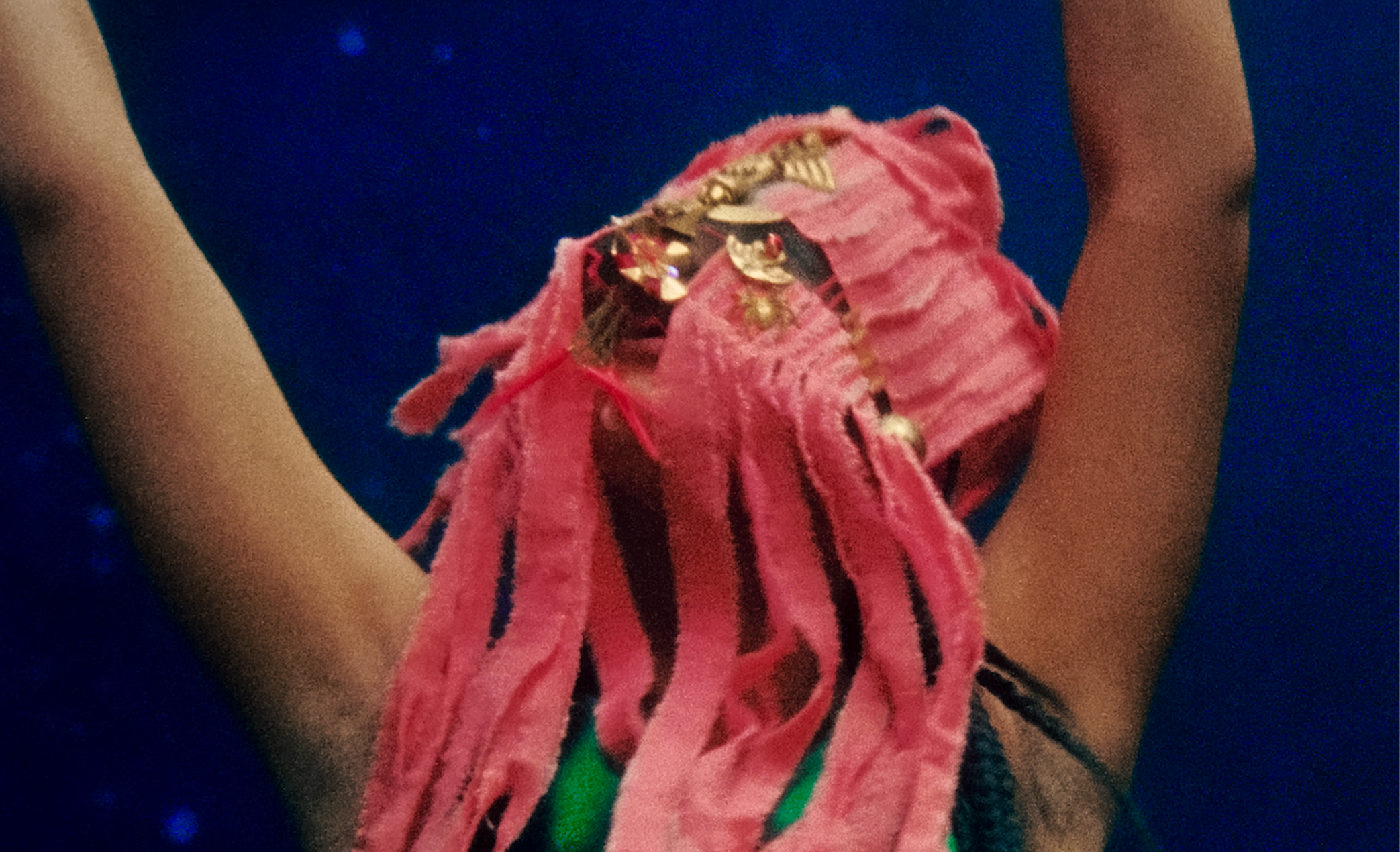 Close-up of a person wearing a pink hoodie with black stripes and a necklace. The background is dark with small blue lights.