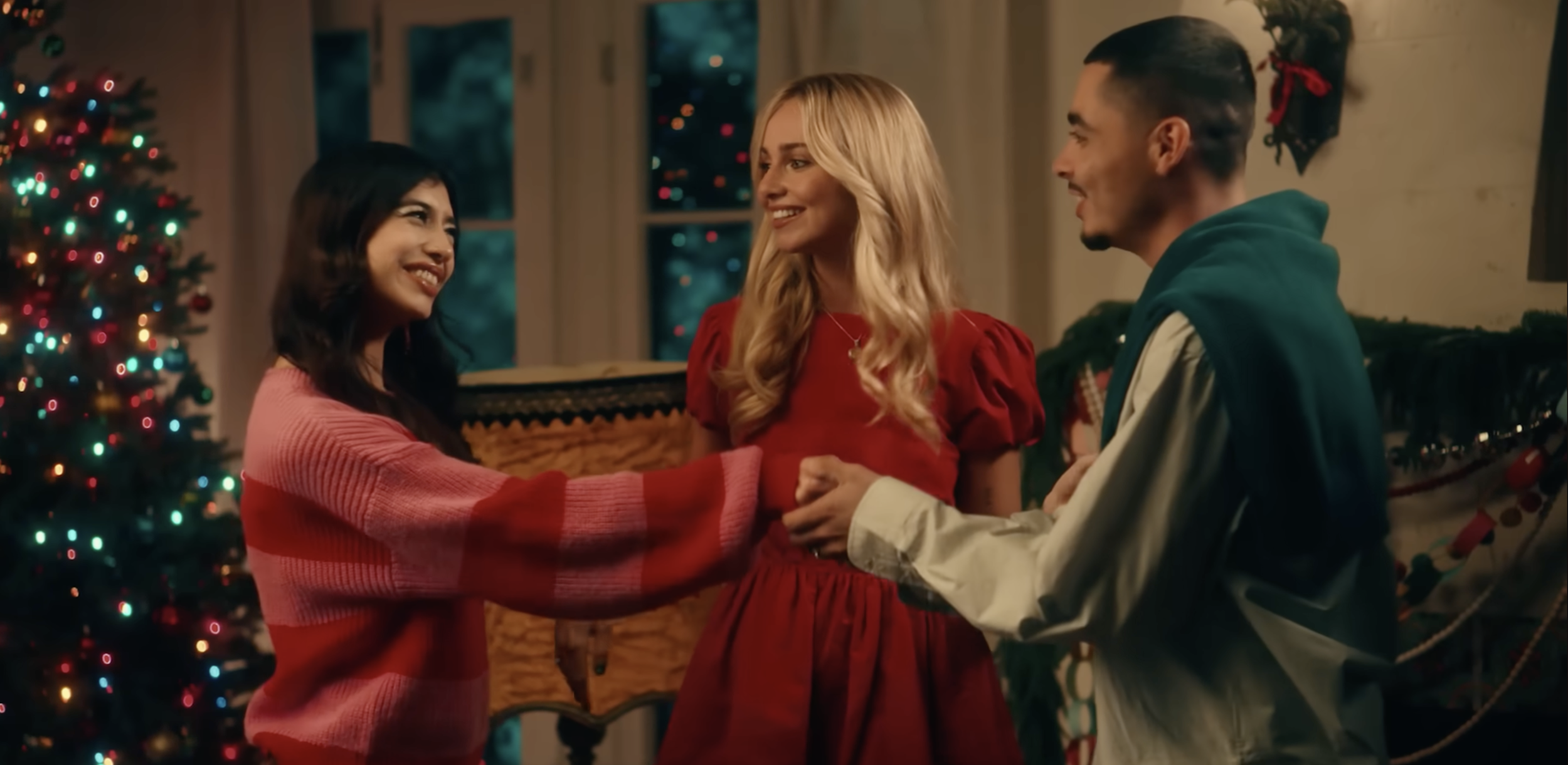 Three people holding hands and smiling, standing near a decorated Christmas tree inside a warmly lit room, with a window showing Christmas lights outside.