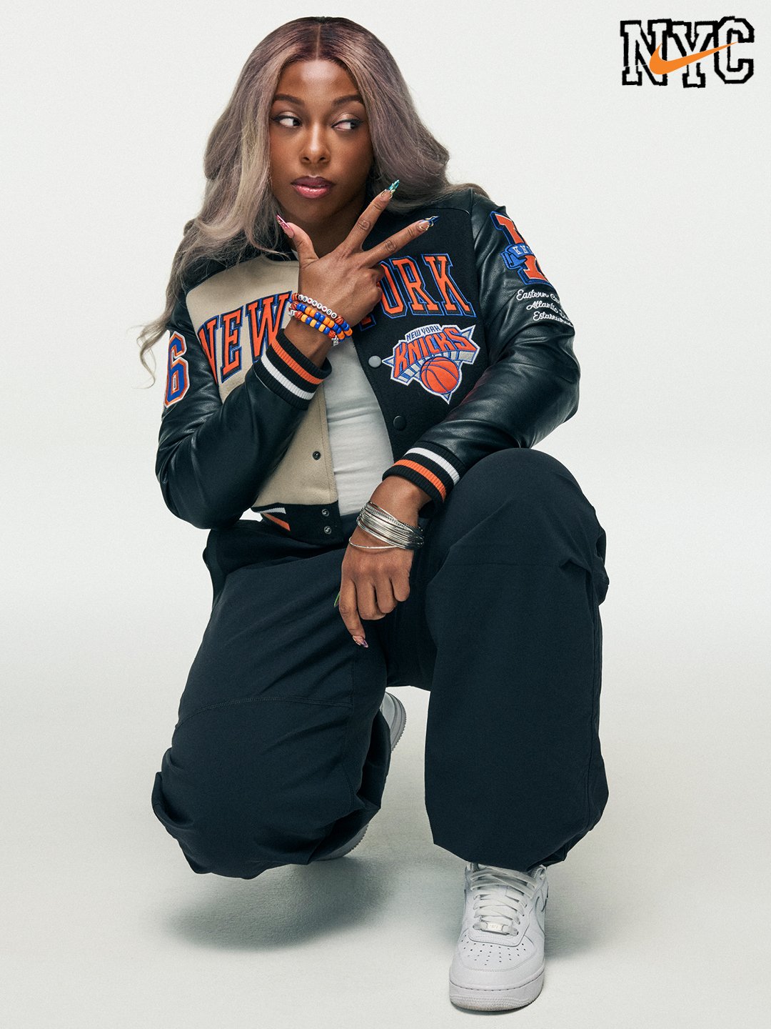 A young woman with dyed gray hair posing in a New York Knicks jacket, black pants, and white sneakers, making a peace sign with her right hand, against a plain backdrop with NYC logo in the top right corner.