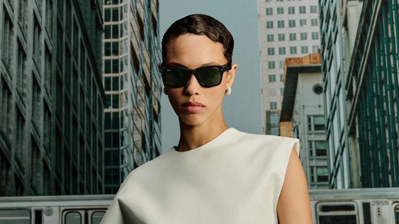 A woman wearing black sunglasses and a white sleeveless top standing in a city street with tall buildings and an elevated train in the background.