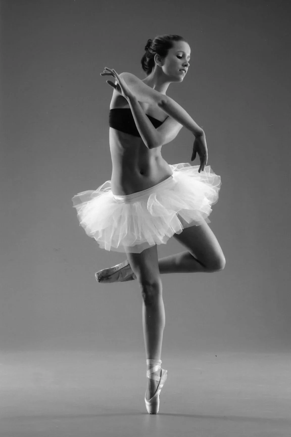 Black and white photo of a ballerina in a tutu, balancing on one foot en pointe with arms crossed in front of her, against a plain background.