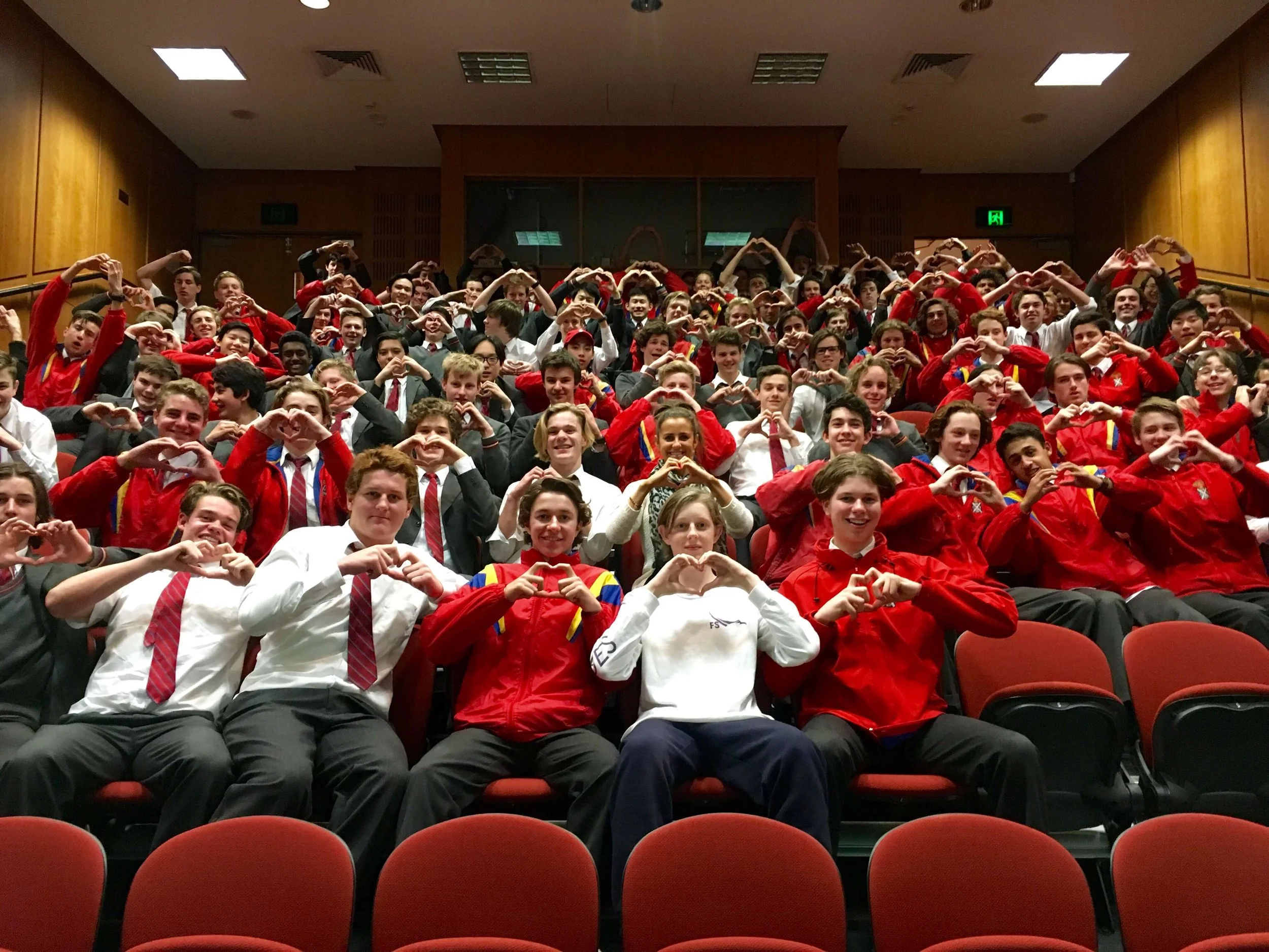 A large group of students, dressed in uniforms and red jackets, sitting in theater seats in an auditorium. Many are making heart shapes with their hands and smiling at the camera.
