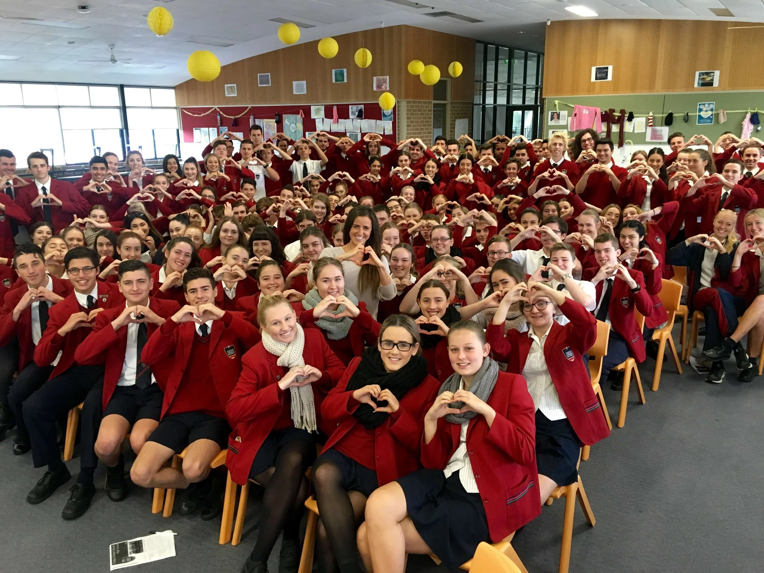 Group of students in a classroom making heart shapes with their hands, dressed in red uniforms, smiling, with colorful decorations hanging from the ceiling.