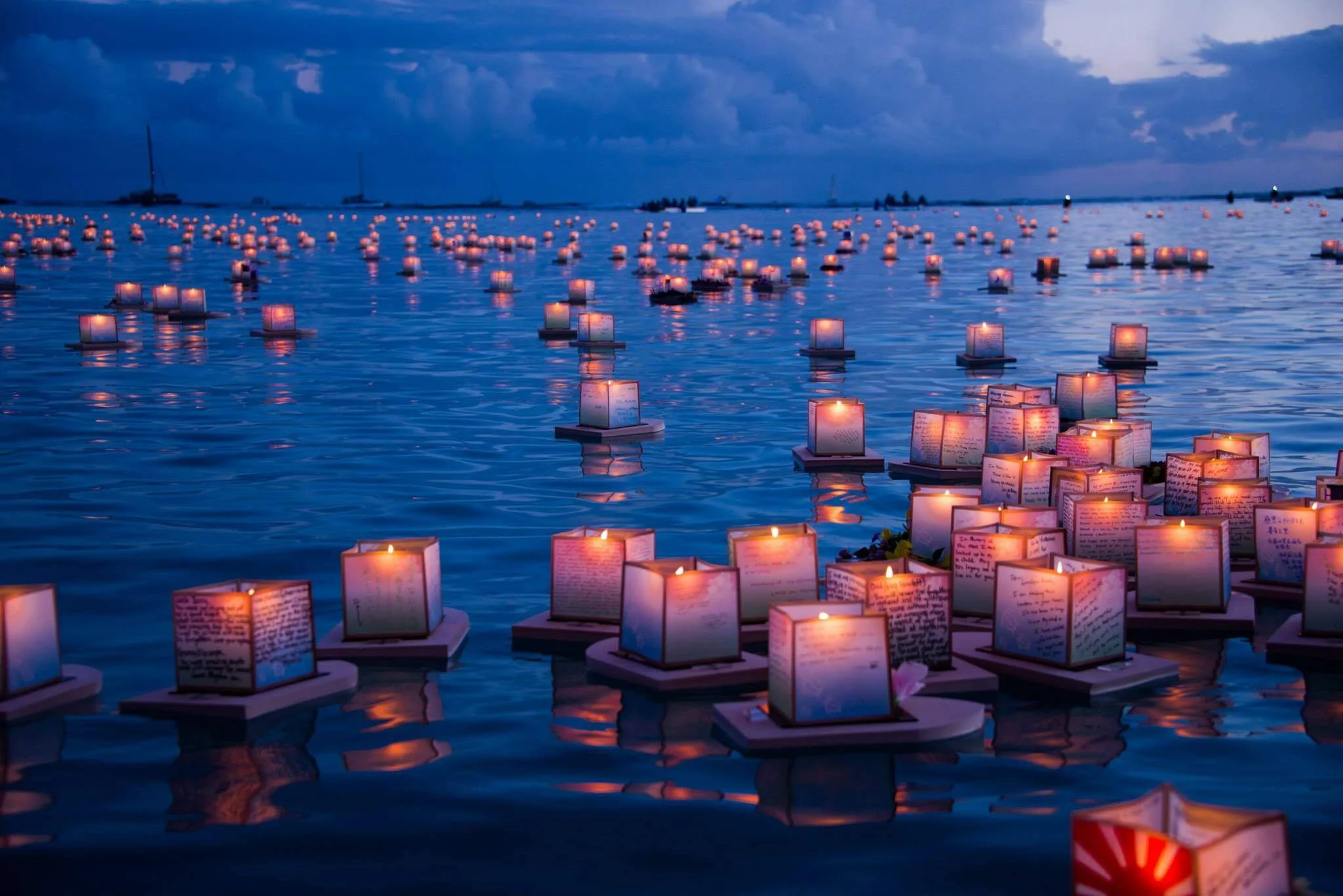 Numerous floating lanterns with lit candles on water, with handwritten notes or messages on each lantern, during twilight or early evening, creating a peaceful and reflective atmosphere.