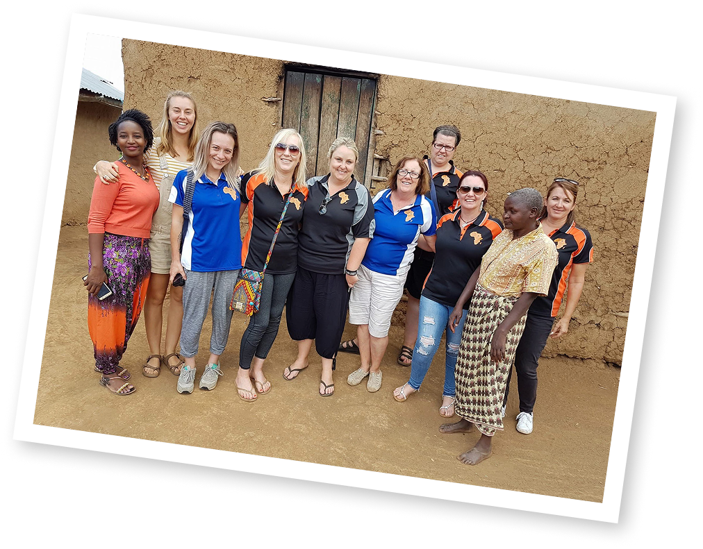 Group of eleven women standing outdoors in front of a mud wall, smiling, some with sunglasses, dressed in casual and athletic clothing.