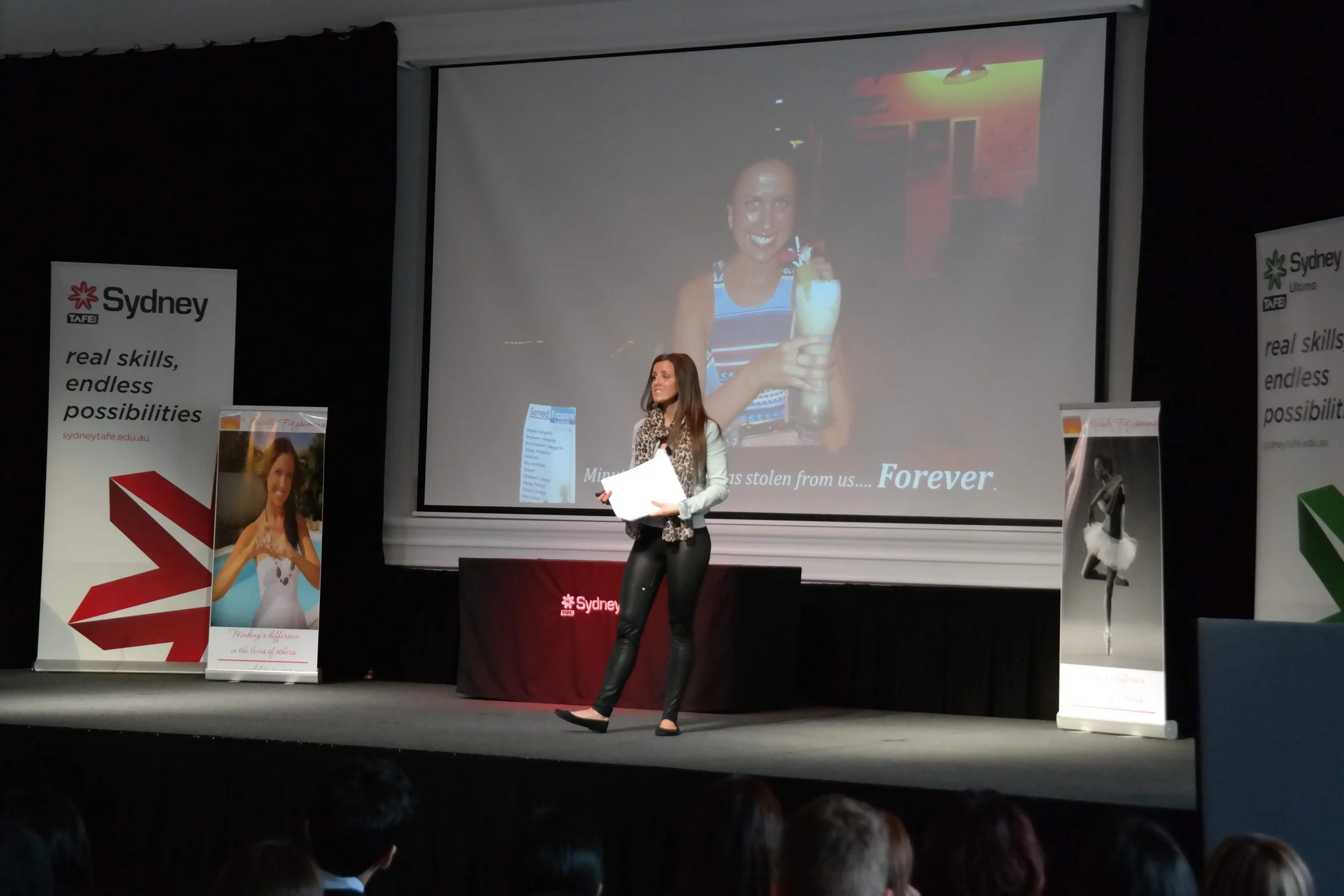 A woman on stage giving a presentation in front of a large projector screen, with banners on either side promoting Sydney TAFE.