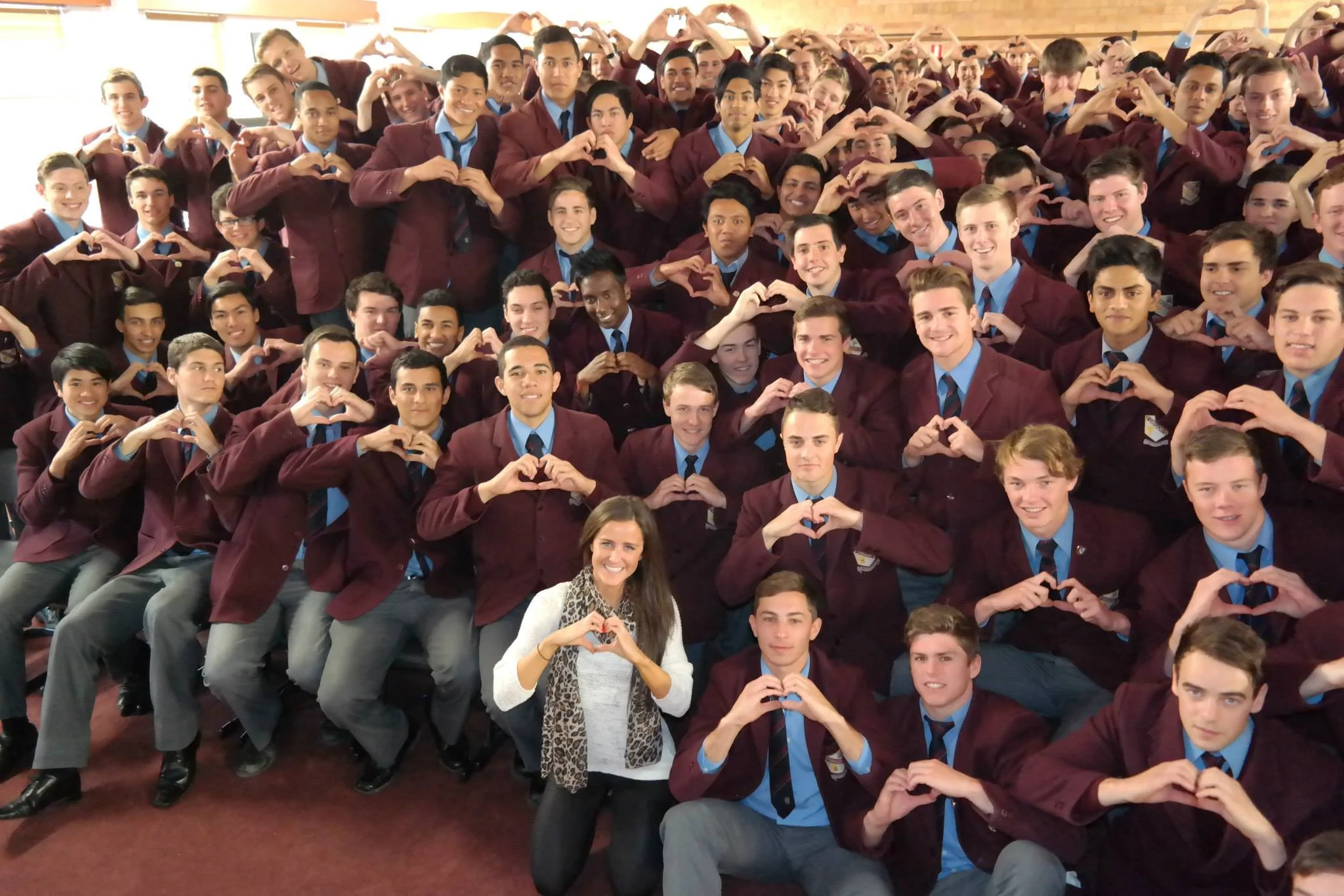 A large group of diverse students in school uniforms, making heart shapes with their hands, gathered in a classroom or assembly hall.