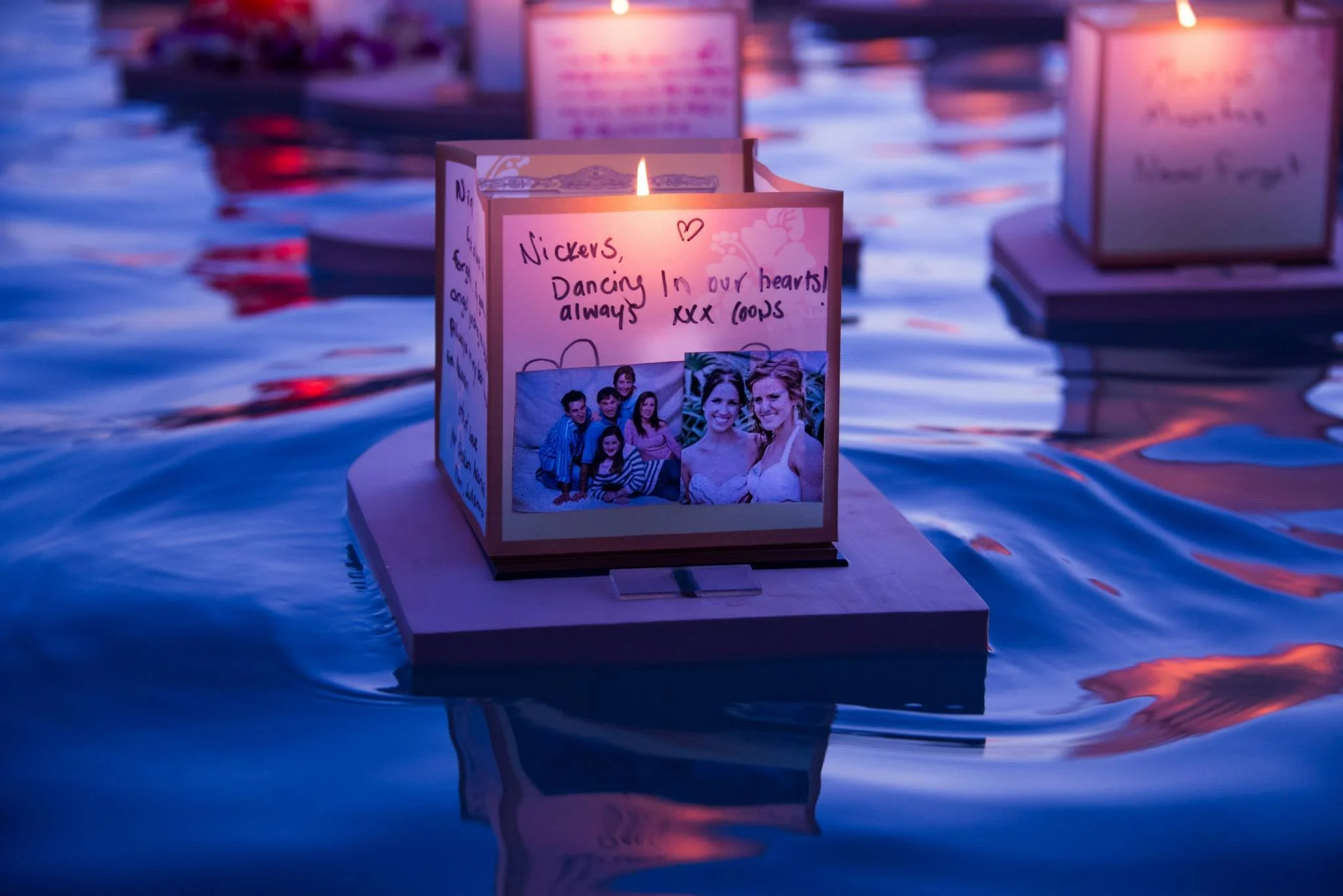 A candle with a handwritten note and a family photo on a table at night, with other candles and signs in the background.
